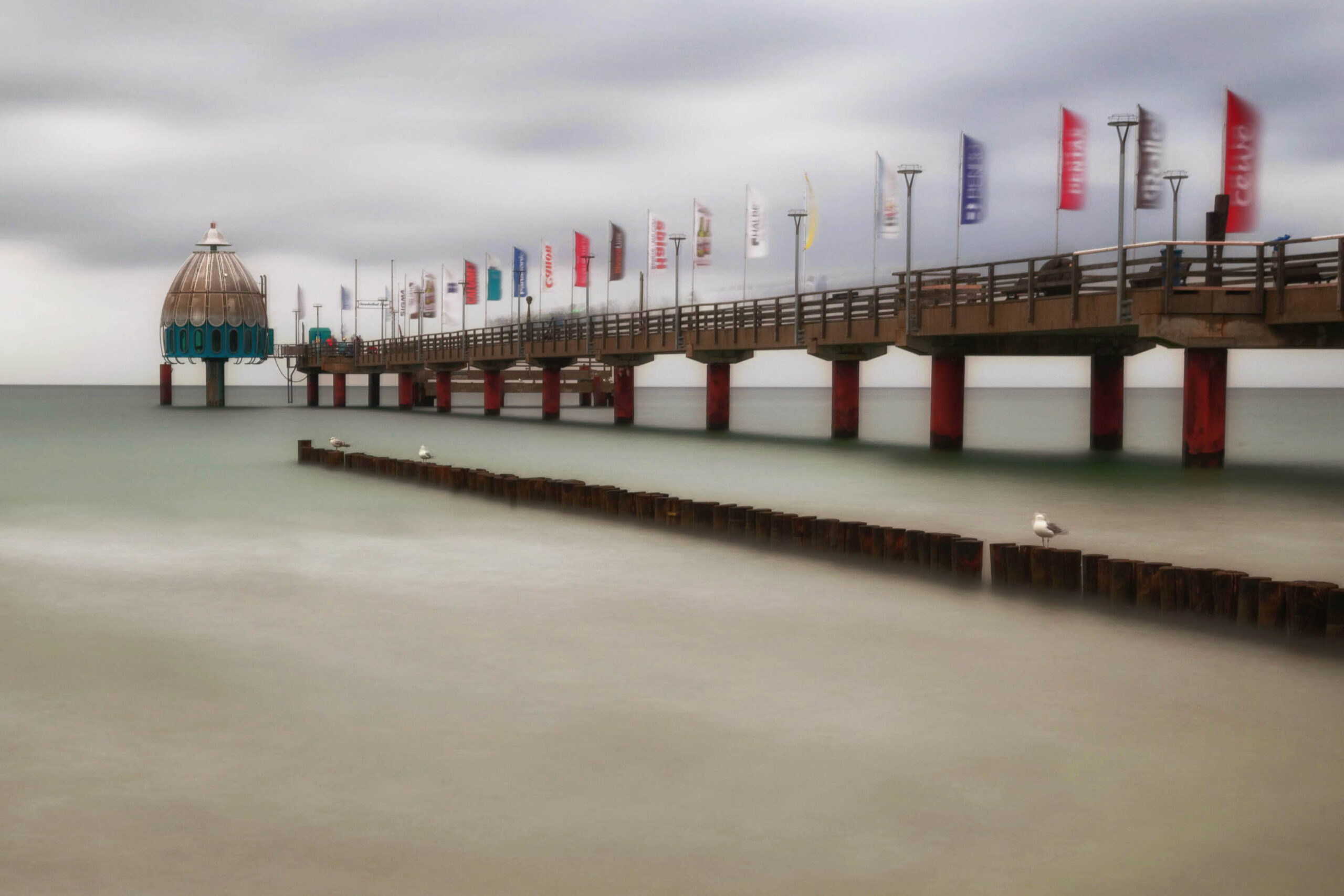 Das Foto zeigt den Steg von Zingst an der Ostsee. Der Steg erstreckt sich weit in den grauen Ozean. Am Ende des Stegs befindet sich ein runder Leuchtturm. Der Himmel ist bedeckt und die See ruhig. Einige Möwen sitzen auf den Holzpfosten des Stegs. Die Aufnahme wurde im Juni 2013 aufgenommen.