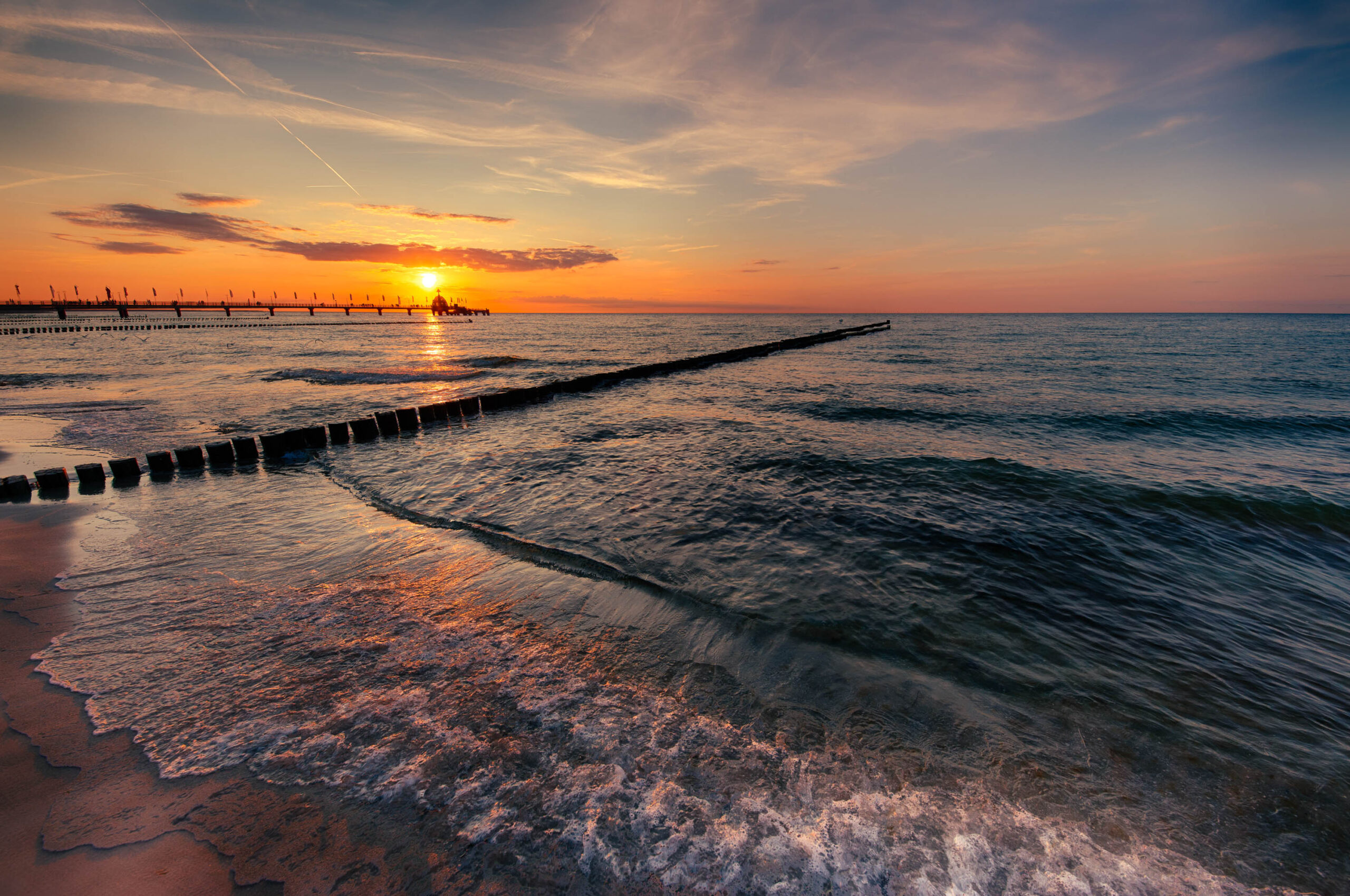 Das Foto zeigt einen malerischen Sonnenuntergang an der Ostsee, genauer gesagt in Zingst. Ein hölzerner Wellenbrecher zieht sich vom Strand ins Meer und wird von den letzten Sonnenstrahlen in ein goldenes Licht getaucht. Das ruhige Wasser reflektiert die warmen Farben des Himmels, was eine friedliche und entspannende Atmosphäre schafft. Der Himmel ist von leichten Wolken durchzogen, die das Licht noch weicher erscheinen lassen. Die Szene vermittelt ein Gefühl von Ruhe und Schönheit der Natur.