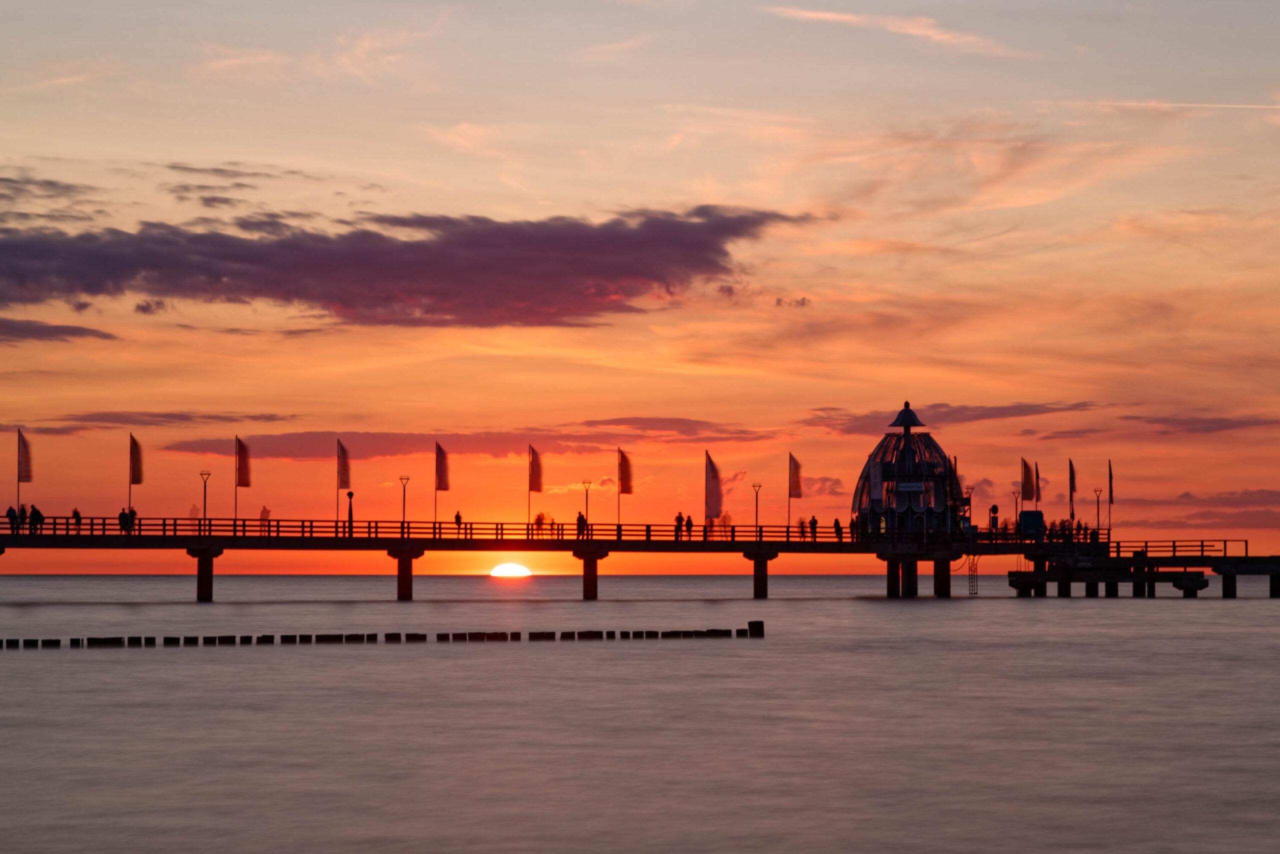 Das Bild zeigt einen Steg, der sich über ruhiges, dunkles Wasser erstreckt. Der Himmel ist in leuchtenden Orange- und Rottönen gefärbt, was auf einen Sonnenuntergang hindeutet. Die Silhouette des Stegs und der darauf stehenden Personen ist deutlich erkennbar. Am Ende des Stegs befindet sich ein markantes, kuppelförmiges Gebäude. Die Wasseroberfläche spiegelt die Farben des Himmels wider und erzeugt eine ruhige und friedliche Atmosphäre. Die Szene vermittelt ein Gefühl von Ruhe und Frieden, typisch für einen Abend am Meer.