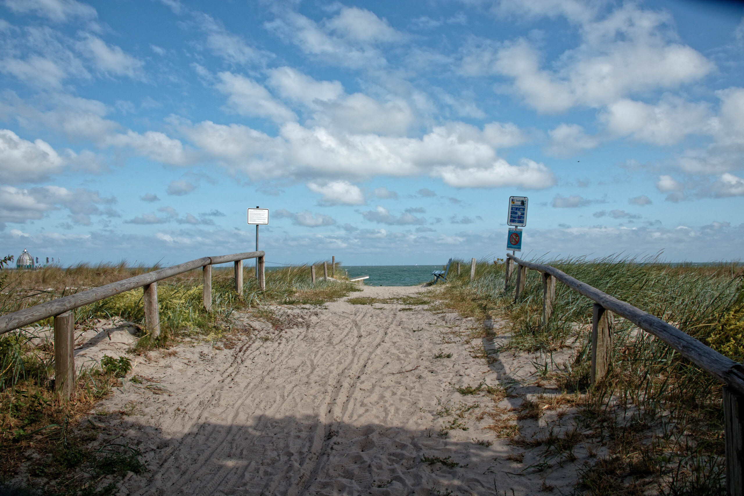 Das Bild zeigt einen Weg, der durch ein Dünengebiet führt und zu einem Strand an der Ostsee führt. Der Weg ist mit Holzpfosten und Geländern gesichert, um den Übergang durch die Dünen zu erleichtern und die Vegetation zu schützen. Der Strand erstreckt sich im Hintergrund, und das Meer ist sichtbar. Der Himmel ist bewölkt, was auf eine leicht wechselhaftes Wetter hindeutet. Die Szene vermittelt ein Gefühl von Ruhe und Erholung.