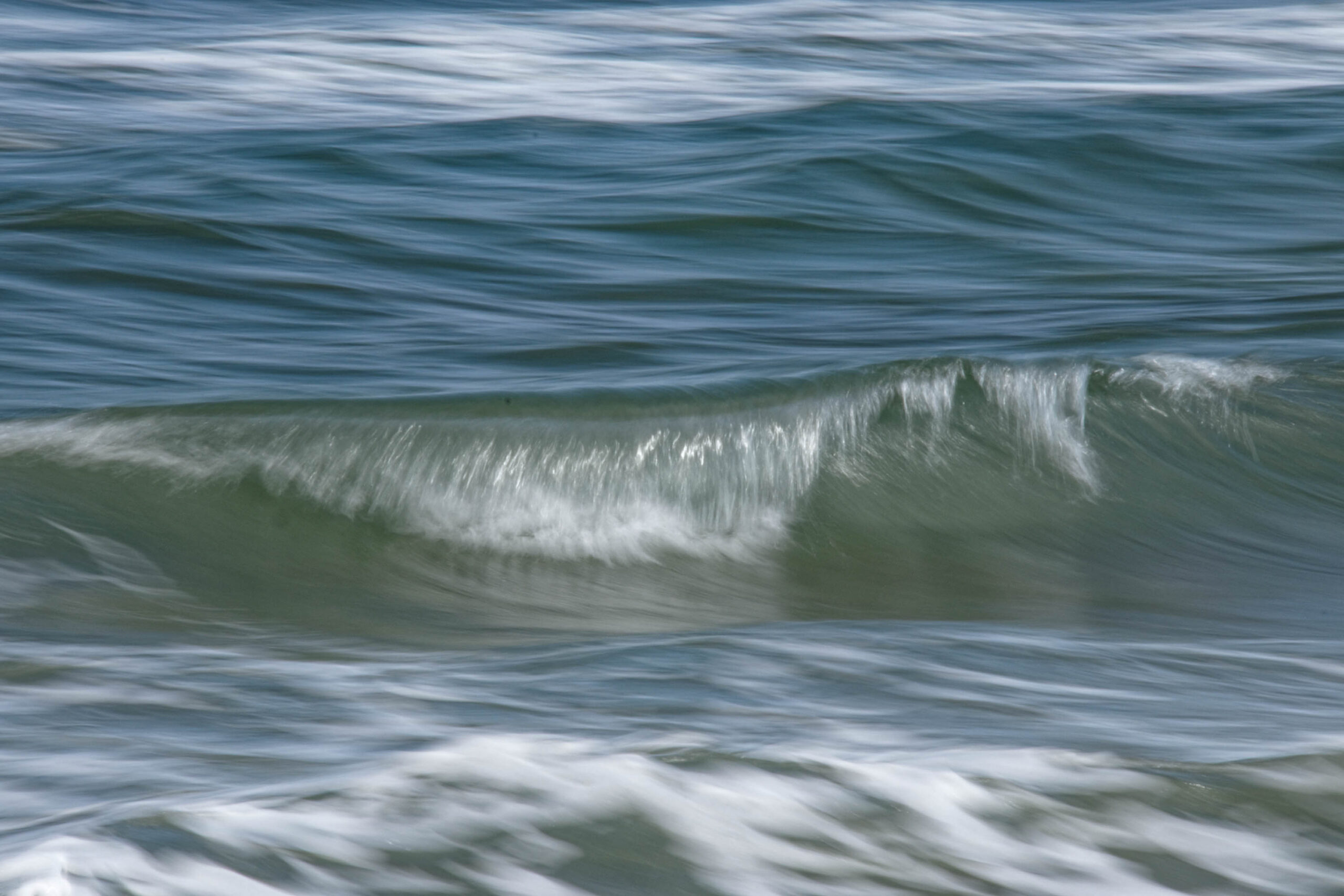 Das Bild zeigt eine Nahaufnahme von Wellen auf dem Strand. Das Wasser ist dunkelgrün und die Wellen brechen mit weißen Schaumkronen. Die Bewegung des Wassers ist durch die lange Belichtungszeit verschwommen dargestellt, was einen ruhigen und fließenden Effekt erzeugt. Der Strand ist nicht sichtbar, da der Fokus ausschließlich auf den Wellen liegt. Die Aufnahme wurde im Juli 2019 am Strand von Zingst, Ostsee, aufgenommen.
