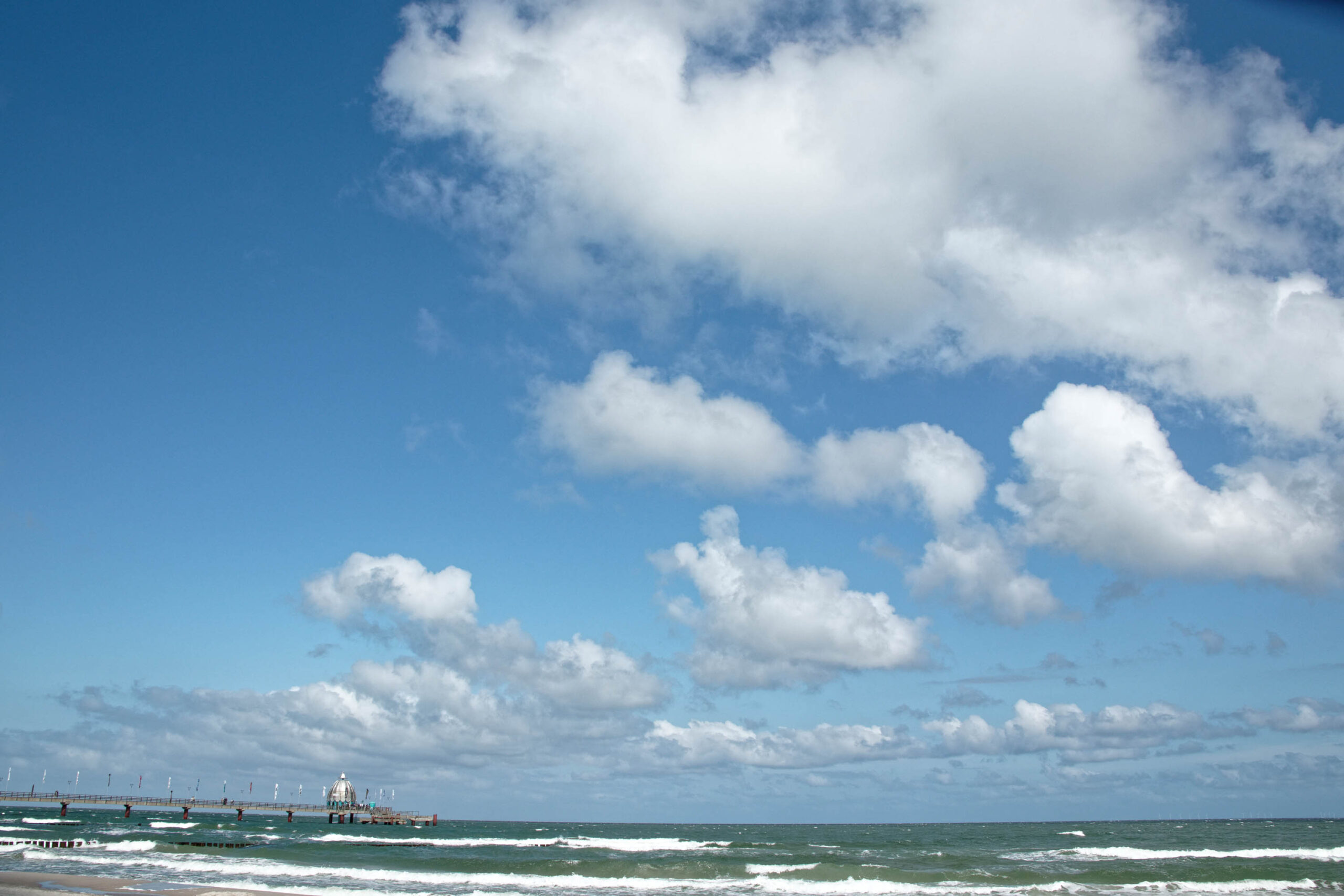 Das Foto zeigt eine malerische Szene vom Strand von Zingst an der Ostsee. Ein hölzerner Steg, der sich in das blaue Meer erstreckt, dominiert den Vordergrund. Das Wasser ist leicht aufgewühlt, mit kleinen Wellen, die an den Strand rollen. Der Himmel ist strahlend blau und von weißen, flockigen Wolken durchzogen. Einige Personen sind auf dem Steg zu sehen, die sich entspannen oder die Aussicht genießen. Die Szene vermittelt ein Gefühl von Ruhe und Erholung.