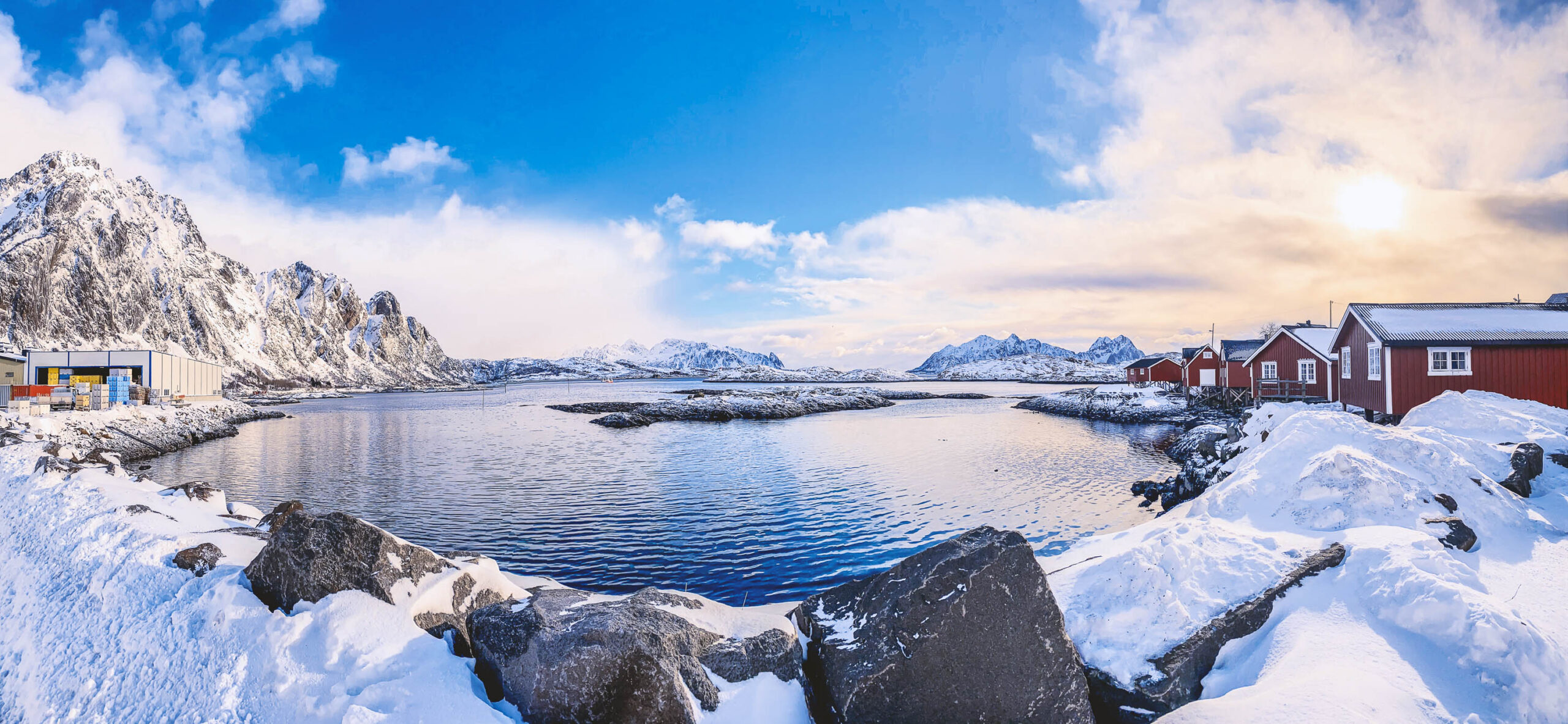 Die Aufnahme fängt die Schönheit der Lofoten-Inseln in Norwegen ein. Im Vordergrund dominieren dunkle Felsen, bedeckt mit einer dicken Schneedecke. Dahinter erstreckt sich ein ruhiges, tiefblaues Meer, das von schneebedeckten Bergen gesäumt wird. Am Ufer stehen traditionelle rote Fischerhütten, die einen charmanten Kontrast zu der rauen Landschaft bilden. Der Himmel ist bewölkt, mit vereinzelten Lichtungen, die einen sanften Schein auf die Szene werfen. Die Aufnahme vermittelt ein Gefühl von Frieden und Erhabenheit.