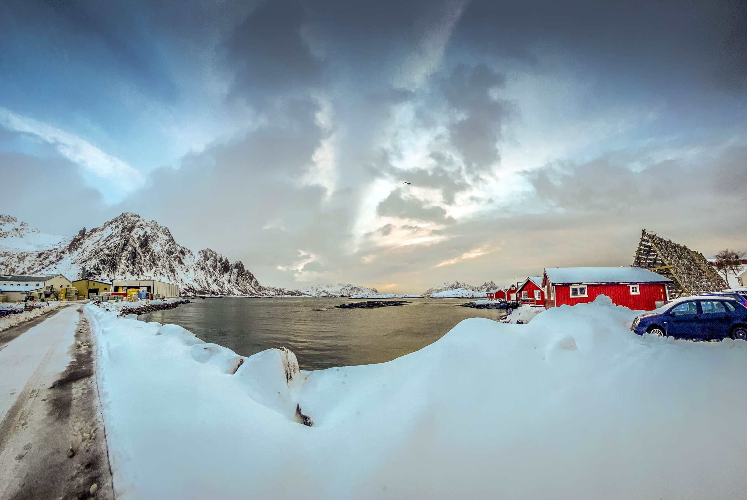 Die Fotografie zeigt eine malerische Winterlandschaft in den Lofoten, Norwegen. Im Vordergrund liegt eine dicke Schneedecke, die einen gewundenen Weg säumt. Im Hintergrund erheben sich schneebedeckte Berge, die den Blick auf eine Reihe von traditionellen roten Fischerhütten am Ufer eines Fjords freigeben. Der Himmel ist bewölkt, mit einem sanften Licht, das die Szene erhellt. Die Farben sind gedämpft, was die kalte und ruhige Atmosphäre der nordnorwegischen Landschaft unterstreicht. Die Szene vermittelt ein Gefühl von Frieden und Abgeschiedenheit.
