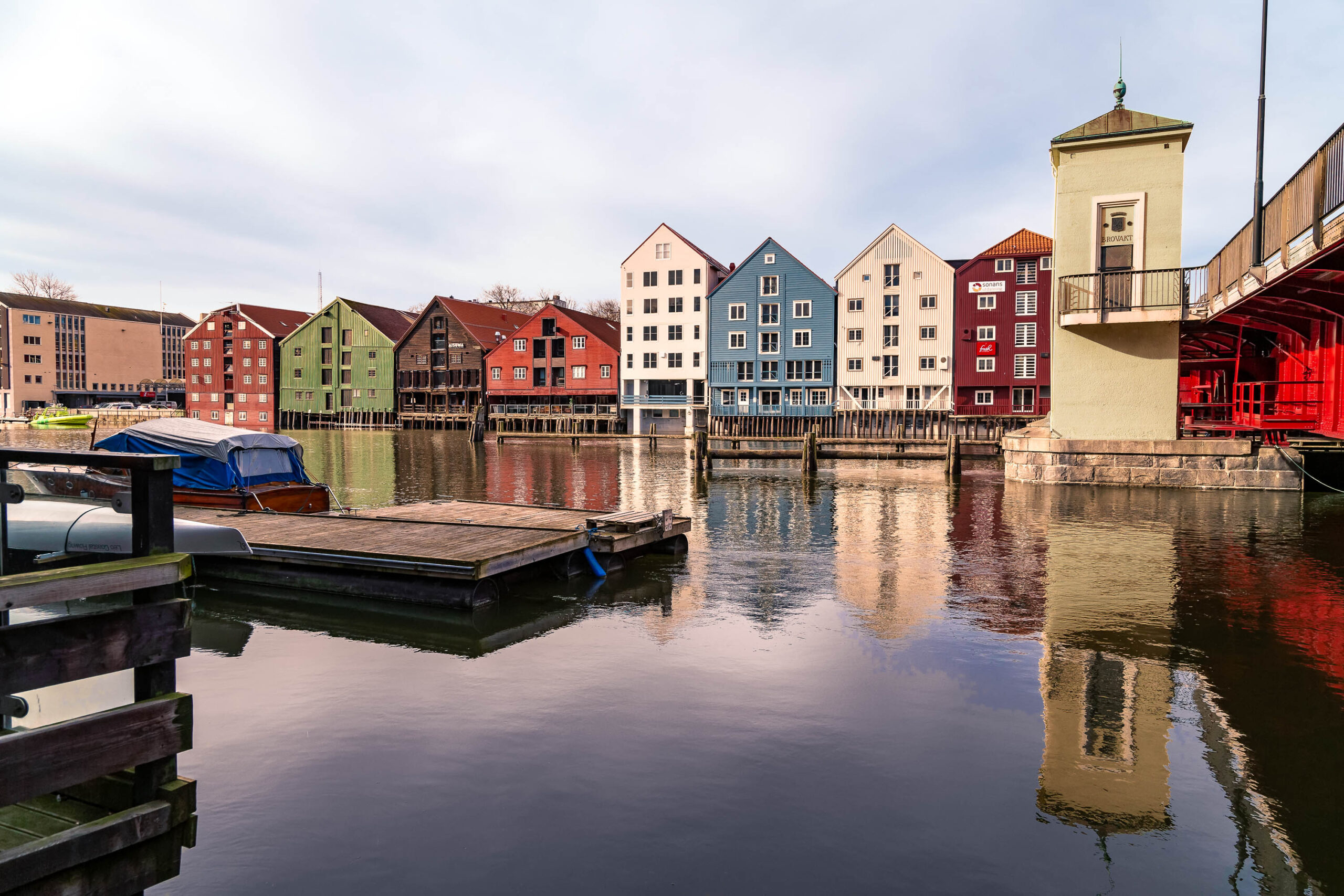 Die Aufnahme zeigt eine malerische Szene entlang des Nidelva-Kanals in Trondheim, Norwegen. Die historischen Gebäude, die sich entlang des Kanals erstrecken, sind in einer Vielzahl von Farben gestrichen, darunter Rot, Grün, Blau und Gelb. Die Gebäude sind typisch für die Architektur Trondheims und zeugen von der reichen Geschichte der Stadt. Das ruhige Wasser des Kanals spiegelt die Gebäude und die umliegende Landschaft wider, was die Szene noch atmosphärischer macht. Eine Holzplattform im Vordergrund verdeckt einen Teil des Wassers, was dem Bild eine interessante Perspektive verleiht. Die Aufnahme wurde im März 2020 aufgenommen, was sich in dem trüben Licht und dem kühlen Aussehen der Szene widerspiegelt.