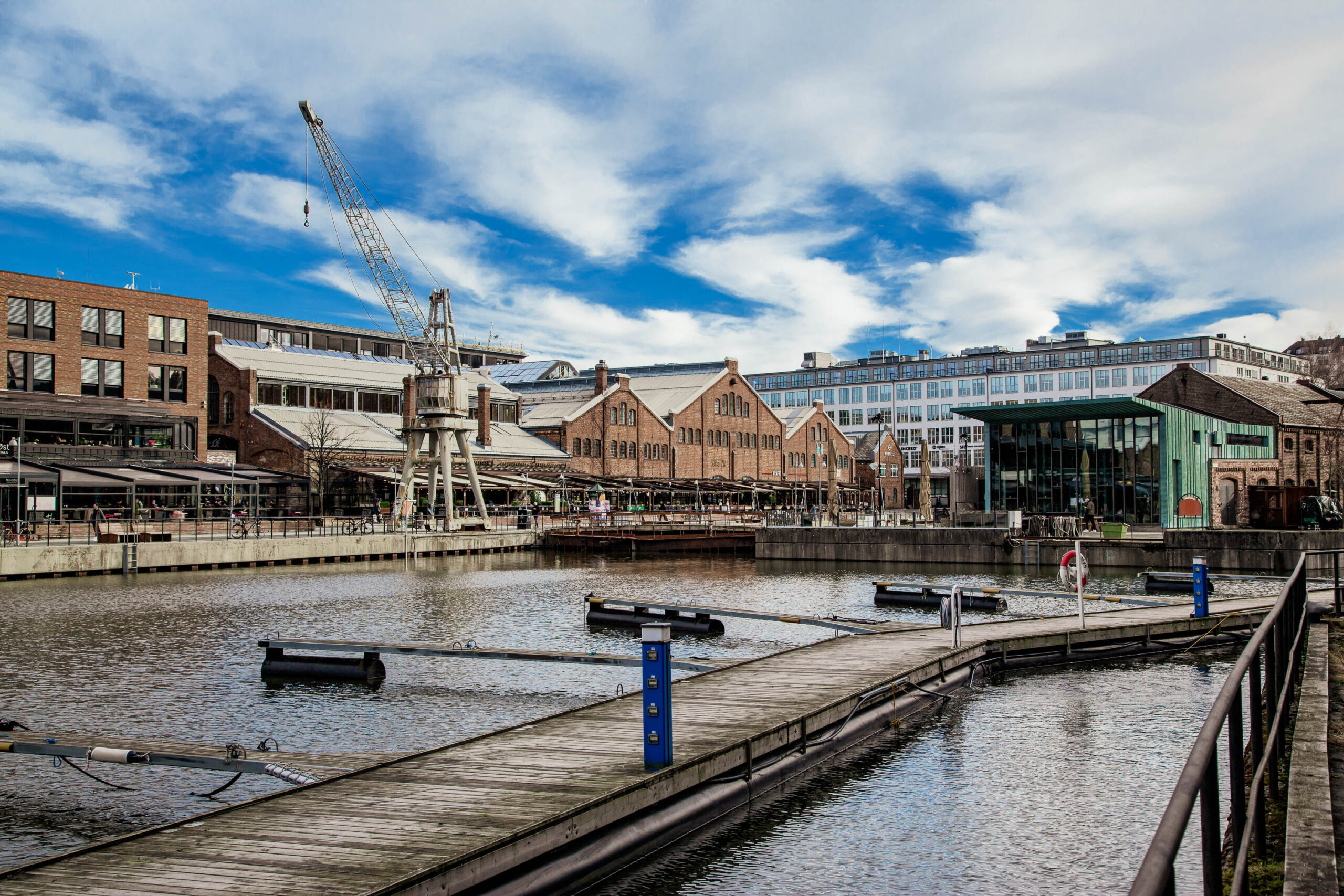 Das Bild zeigt eine malerische Hafenszene in Trondheim, Norwegen. Ein Holzsteg erstreckt sich über das Wasser und führt zu einer Reihe von historischen Lagerhäusern, die sich entlang des Hafens befinden. Die Gebäude sind in verschiedenen architektonischen Stilen gehalten, darunter Backstein und Fachwerk. Der Himmel ist bewölkt, aber es gibt Licht, das auf das Wasser und die Gebäude fällt. Im Hintergrund sind einige moderne Gebäude zu sehen, die einen Kontrast zu den historischen Lagerhäusern bilden. Die Szene vermittelt ein Gefühl von Geschichte, Charme und Ruhe.