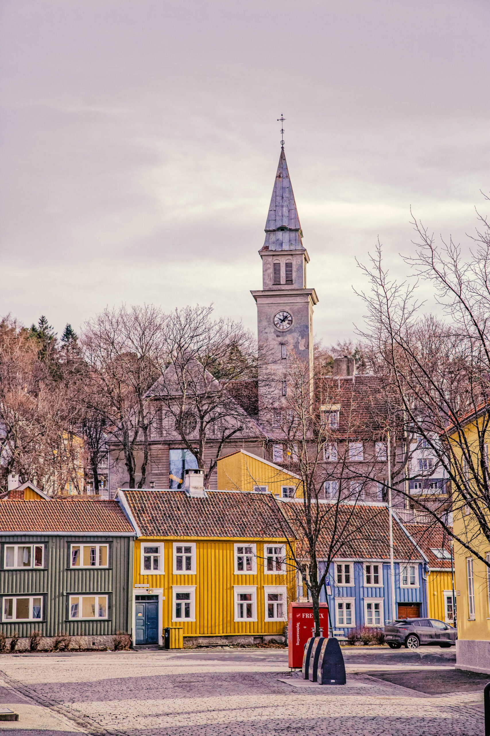 Die Aufnahme zeigt eine typische Straßenszene in Trondheim, Norwegen. Im Vordergrund befindet sich ein gepflasterter Platz, der leicht mit Schnee bedeckt ist. Rechts und links des Platzes reihen sich farbenfrohe, traditionelle Holzhäuser aneinander. Die Häuser sind in verschiedenen Farben gestrichen, darunter Grün, Blau, Gelb und Rot, und verfügen über charakteristische, kleinen Fenster und steile Dächer. Im Hintergrund erhebt sich der Turm der Stiftskirche (Nidarosdomen), ein bedeutendes historisches Bauwerk. Der Himmel ist bedeckt und erzeugt eine ruhige, fast melancholische Stimmung. Die Szene vermittelt ein Gefühl von Tradition und Geschichte.