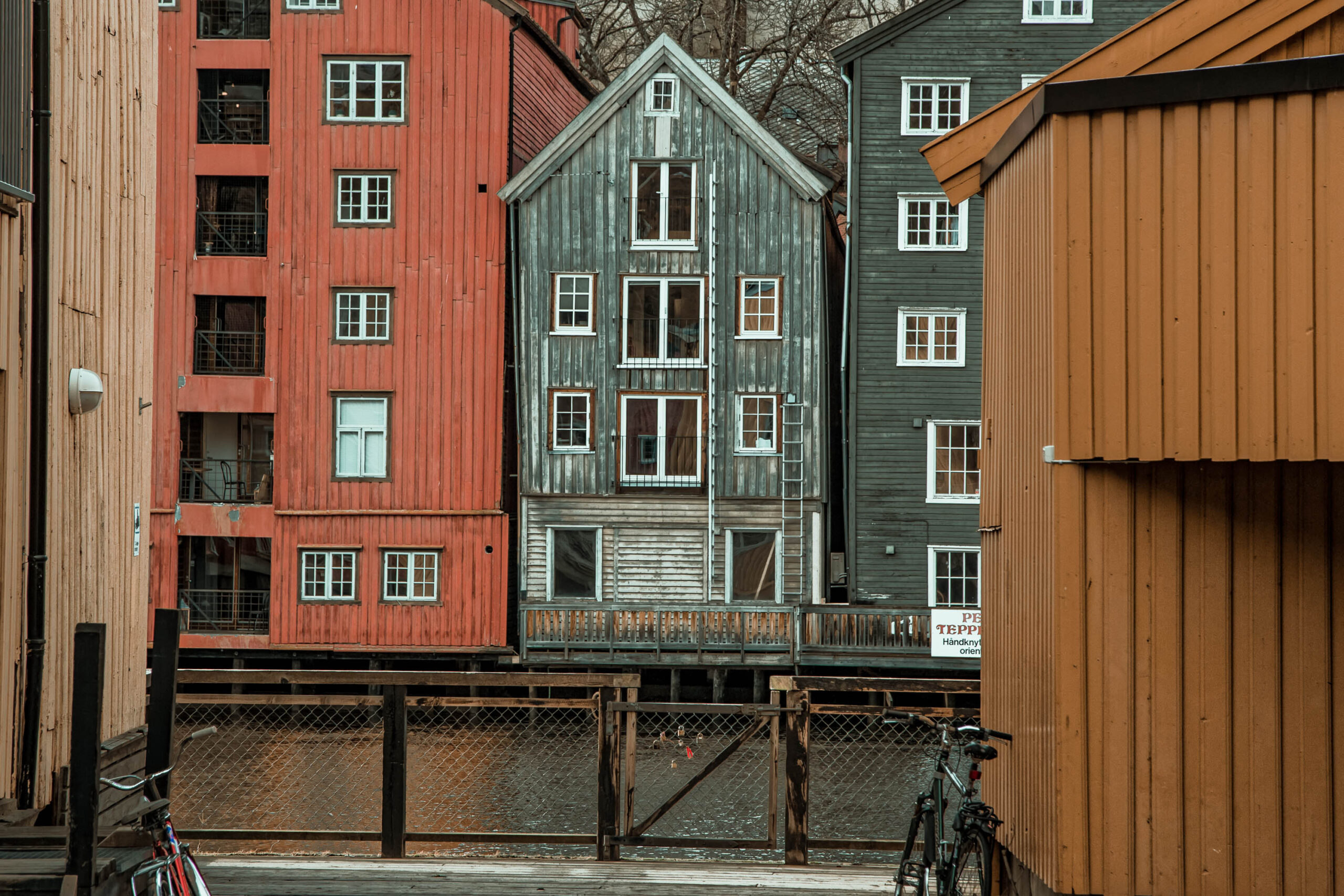 Die Aufnahme zeigt eine Reihe historischer Lagerhäuser in Trondheim, Norwegen. Die Gebäude sind aus Holz und haben unterschiedliche Fassadenfarben: Rot, Grün und Blau. Sie stehen auf Stelzen über einem Kanal. Die Fenster sind mit unterschiedlichen Farben und Designs versehen. Im Vordergrund sind Fahrräder geparkt, was auf eine lebendige Umgebung hindeutet. Die Aufnahme wurde im März 2020 aufgenommen und fängt die einzigartige Architektur und Atmosphäre der Stadt ein.