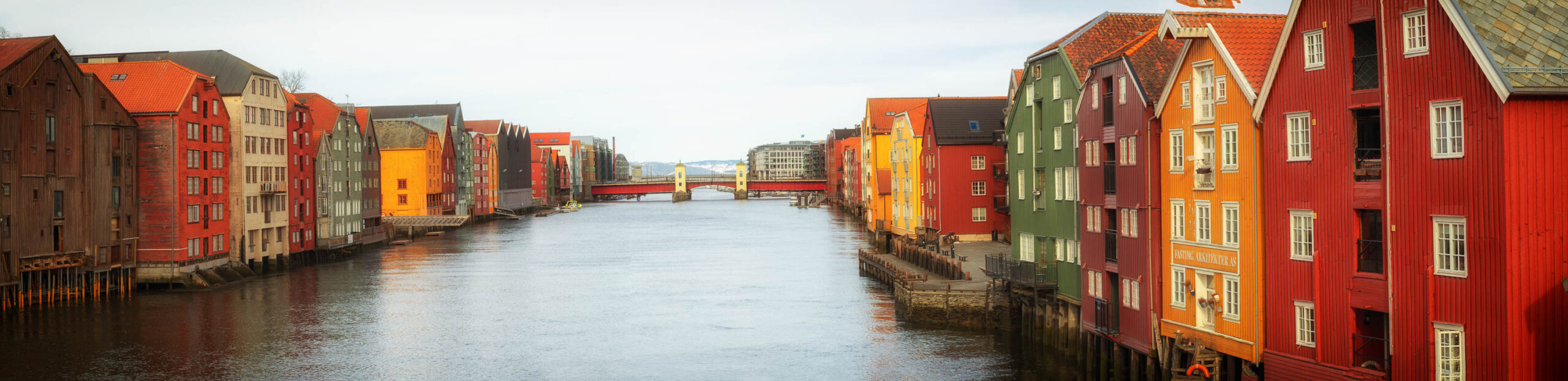 Das Bild zeigt eine malerische Szene in Trondheim, Norwegen. Der Blick fällt auf einen Fluss, der von historischen Gebäuden gesäumt ist, die in leuchtenden Farben gestrichen sind – hauptsächlich Rot, Grün und Orange. Die Gebäude sind typisch für die norwegische Architektur und weisen steile Dächer und hölzerne Fassaden auf. Im Hintergrund ist eine Brücke zu sehen, die den Fluss überspannt. Das Wasser des Flusses ist ruhig und spiegelt die Gebäude und den Himmel wider. Die Szene vermittelt ein Gefühl von Geschichte, Charme und Ruhe.