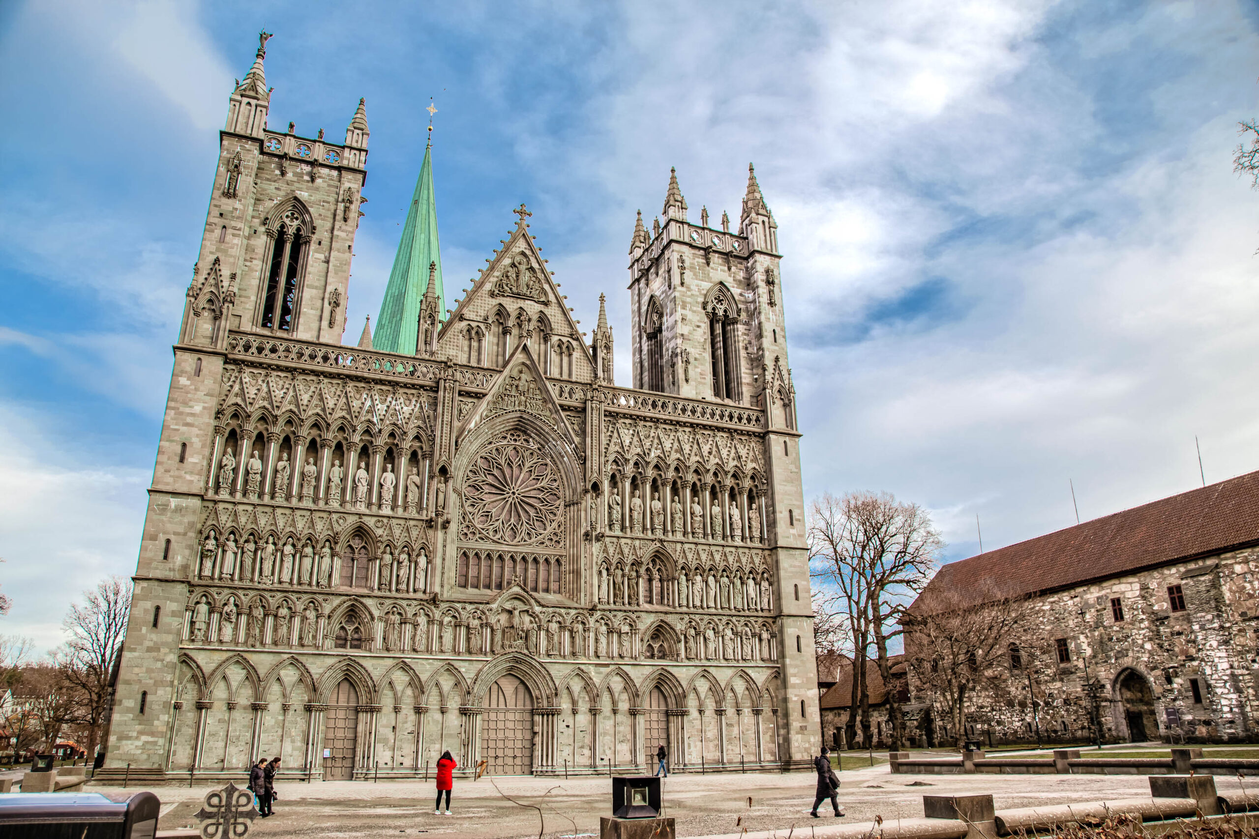 Die Aufnahme zeigt die Nidaros-Kathedrale in Trondheim, Norwegen, im März 2020. Die Kathedrale ist ein beeindruckendes Beispiel gotischer Architektur mit ihren hohen Türmen, den filigranen Steinmetzarbeiten und dem großen Rosfenster. Vor der Kathedrale befindet sich ein großer Platz, auf dem sich einige Personen aufhalten. Im Hintergrund ist ein Gebäude mit einem roten Ziegeldach zu sehen. Der Himmel ist bewölkt, aber es gibt Licht, das die Fassade der Kathedrale beleuchtet.