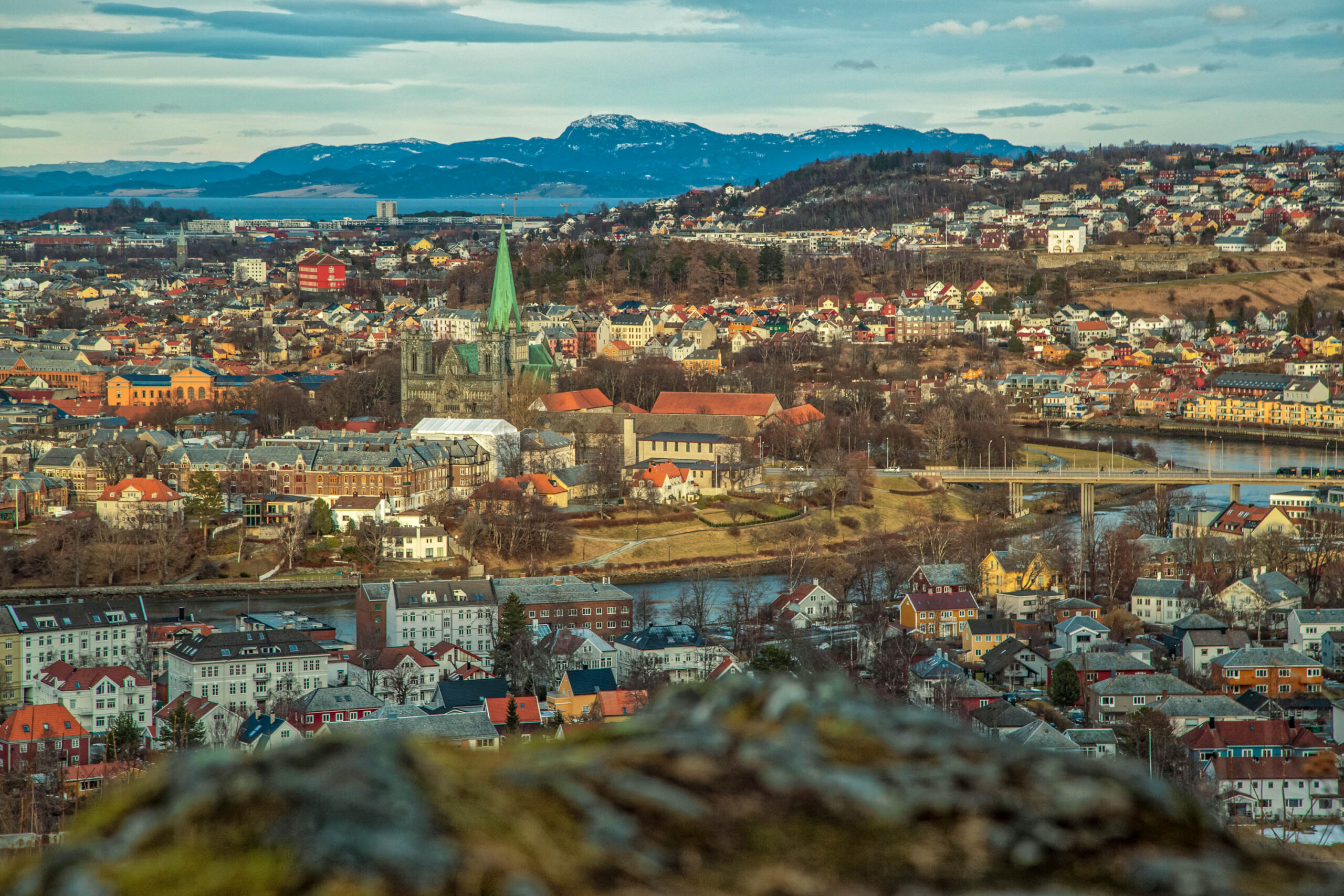 Die Aufnahme zeigt eine weitläufige Panoramaaufnahme von Trondheim, Norwegen, aufgenommen im März 2020. Die Stadt breitet sich über eine hügelige Landschaft aus, wobei die roten Dächer der Häuser einen dominanten visuellen Eindruck hinterlassen. Im Hintergrund erheben sich schneebedeckte Berge, die dem Bild eine dramatische Tiefe verleihen. Der Himmel ist bewölkt, was die Szene in ein gedämpftes Licht taucht. Im Vordergrund ist eine dichte Vegetation zu sehen, die den Blick auf die Stadt teilweise verdeckt. Die Perspektive ist von einem erhöhten Standpunkt aus aufgenommen, was einen umfassenden Überblick über die Stadt und ihre Umgebung ermöglicht.