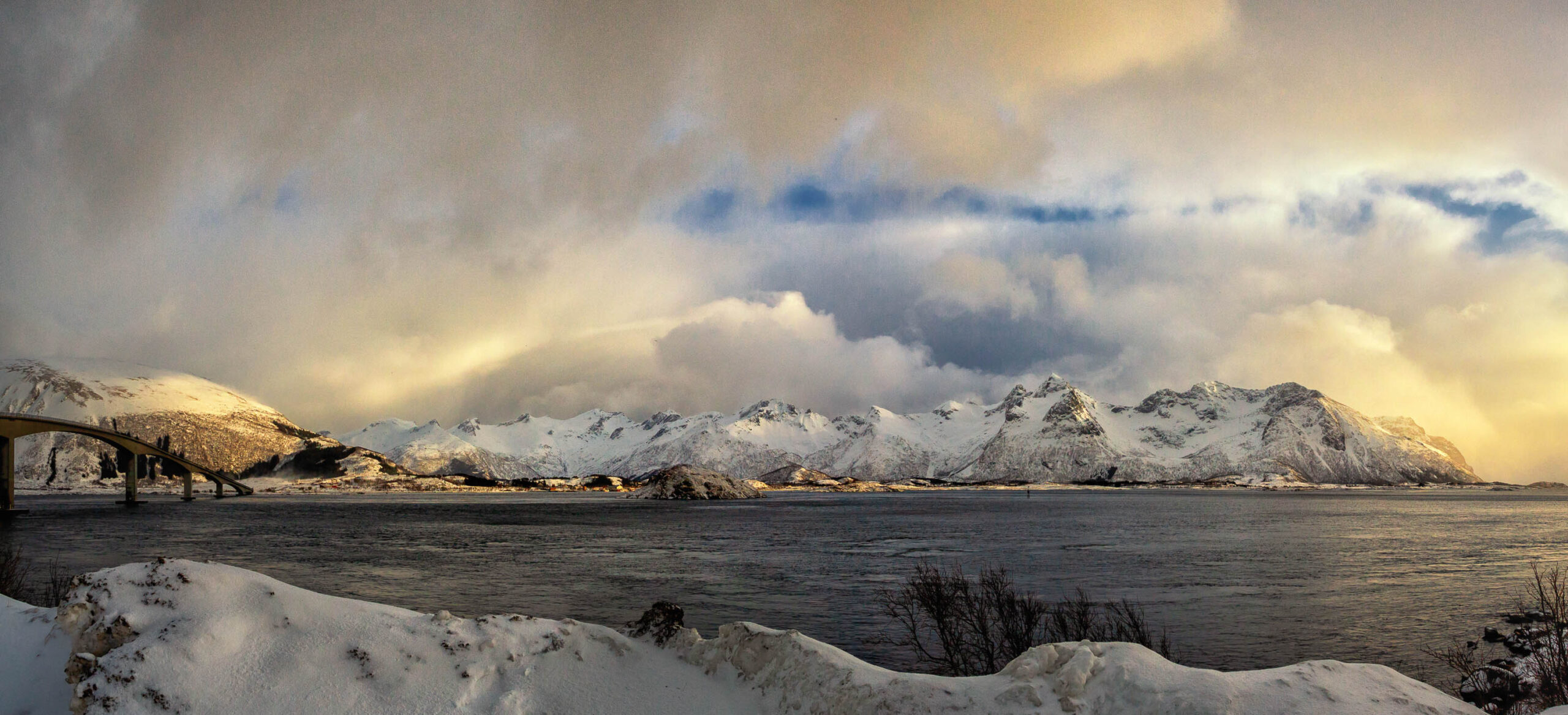 Die Aufnahme zeigt eine typische Lofoten-Landschaft im März 2020. Die schneebedeckten Berge erheben sich majestätisch aus dem dunklen Fjord. Ein Teil einer Brücke ist im Vordergrund links zu sehen. Der Himmel ist bedeckt, aber Lichtstrahlen durchbrechen die Wolken und beleuchten die Szene. Die Farben sind gedämpft, mit einem starken Kontrast zwischen dem dunklen Wasser und den hellen Schneebedeckungen.