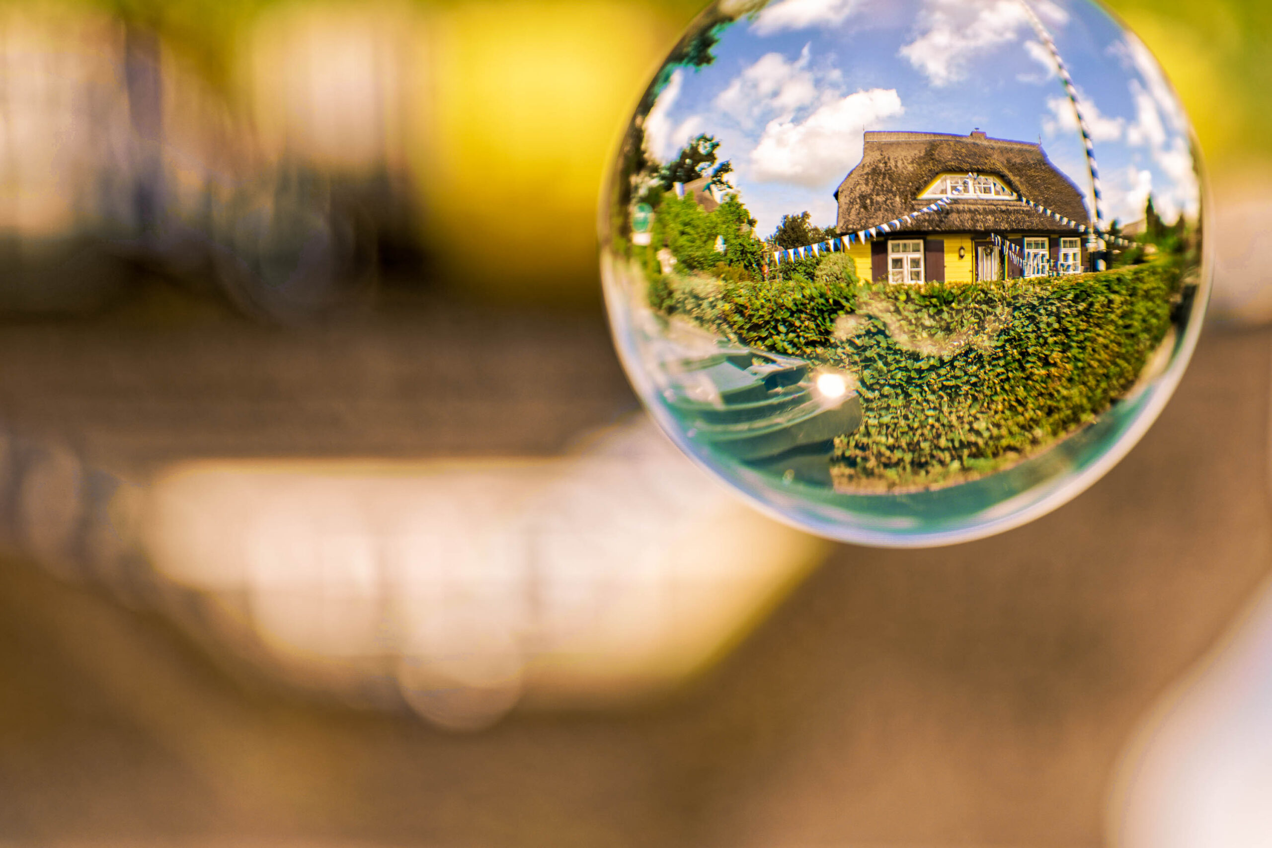 Das Bild zeigt eine Seifenblase, die das Spiegelbild einer traditionellen norddeutschen Fachwerkkulge einfängt. Die Kulge ist mit einem Strohdach gedeckt und verfügt über mehrere Fenster. Die Umgebung ist von dichtem Grün bedeckt, das die Kulge umgibt. Der Himmel ist bewölkt, was dem Bild eine ruhige und friedliche Atmosphäre verleiht. Die Aufnahme wurde während einer Fotosafari im Juli 2020 auf Fischland-Darß-Zingst gemacht.
