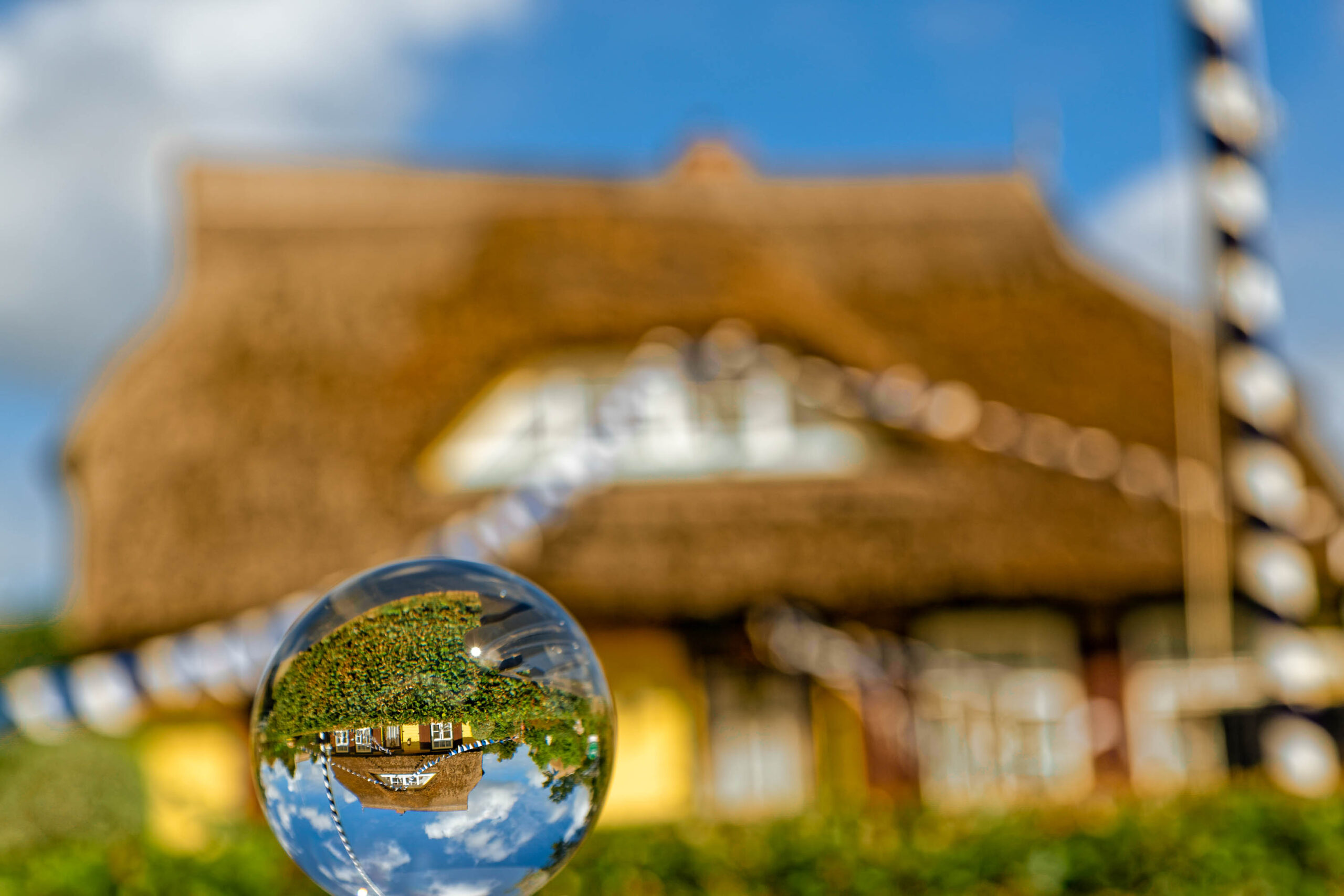 Das Bild zeigt eine ungewöhnliche Perspektive auf ein traditionelles Strohhaus in Fischland-Darss-Zingst. Die Szene wird durch eine sogenannte 'Christbaumkugel' (engl. 'snow globe') reflektiert, was eine verzerrte und verspielte Darstellung der Umgebung erzeugt. Im Vordergrund befindet sich das Gras, das die Kugel hält. Im Hintergrund ist das Strohhaus mit seinem charakteristischen Strohdach zu sehen. Der Himmel ist blau mit vereinzelten Wolken. Die Aufnahme wurde im Juli 2020 während eines Fotosafari gemacht.