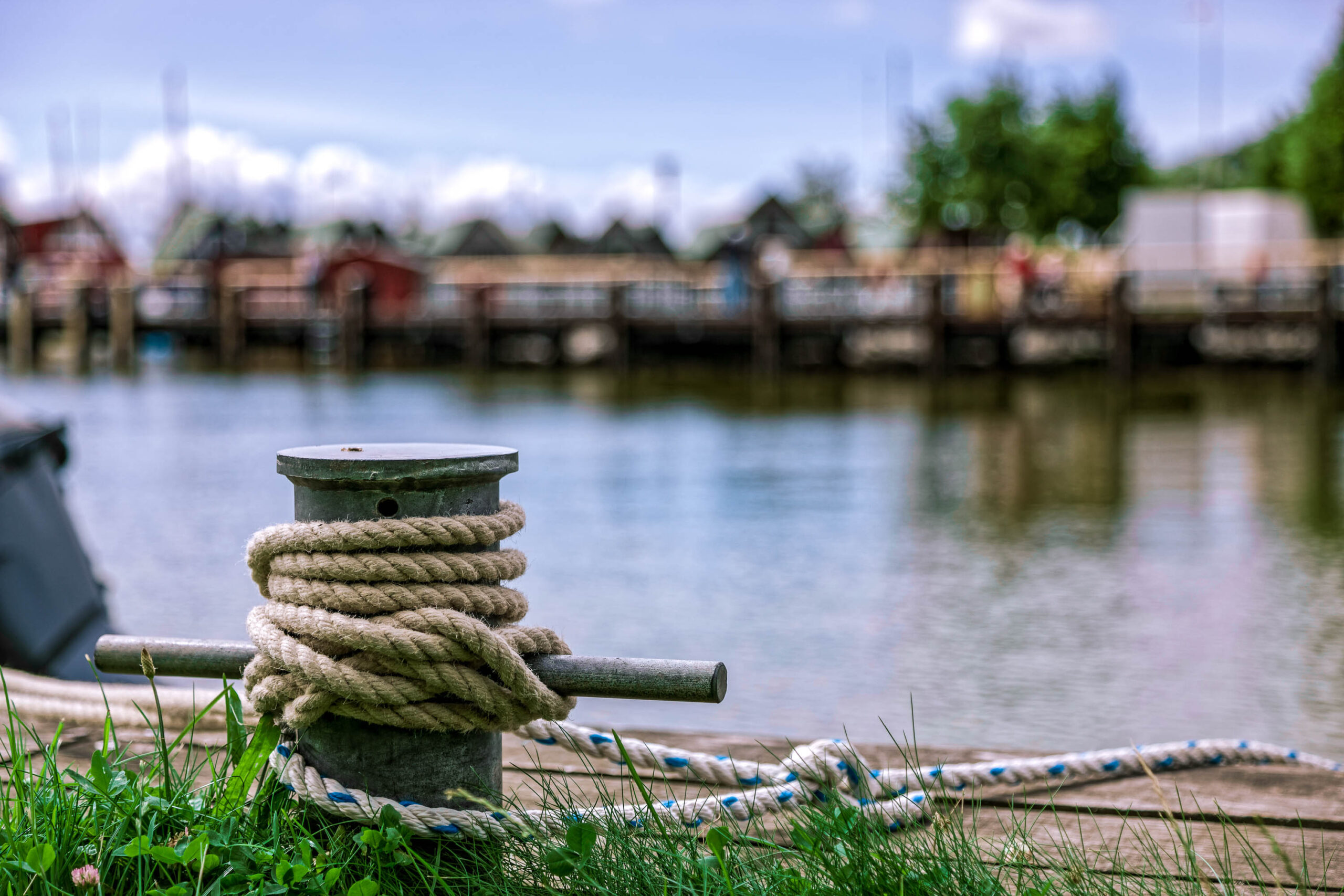 Das Bild zeigt einen Anlegepfahl, an dem eine dicke, braune Schiffslinie festgemacht ist. Der Pfahl ist aus Metall und ragt aus dem Gras heraus. Im Hintergrund ist ein See oder Fluss zu sehen, dessen Oberfläche leicht wellt ist. Am Ufer stehen mehrere Gebäude, die vermutlich Häuser oder Fischerhütten sind. Die Gebäude sind in verschiedenen Farben gestrichen und wirken rustikal. Das Bild ist in einem weichen, verträumten Stil gehalten, mit einem Fokus auf die Textur des Grases und der Schiffslinie.