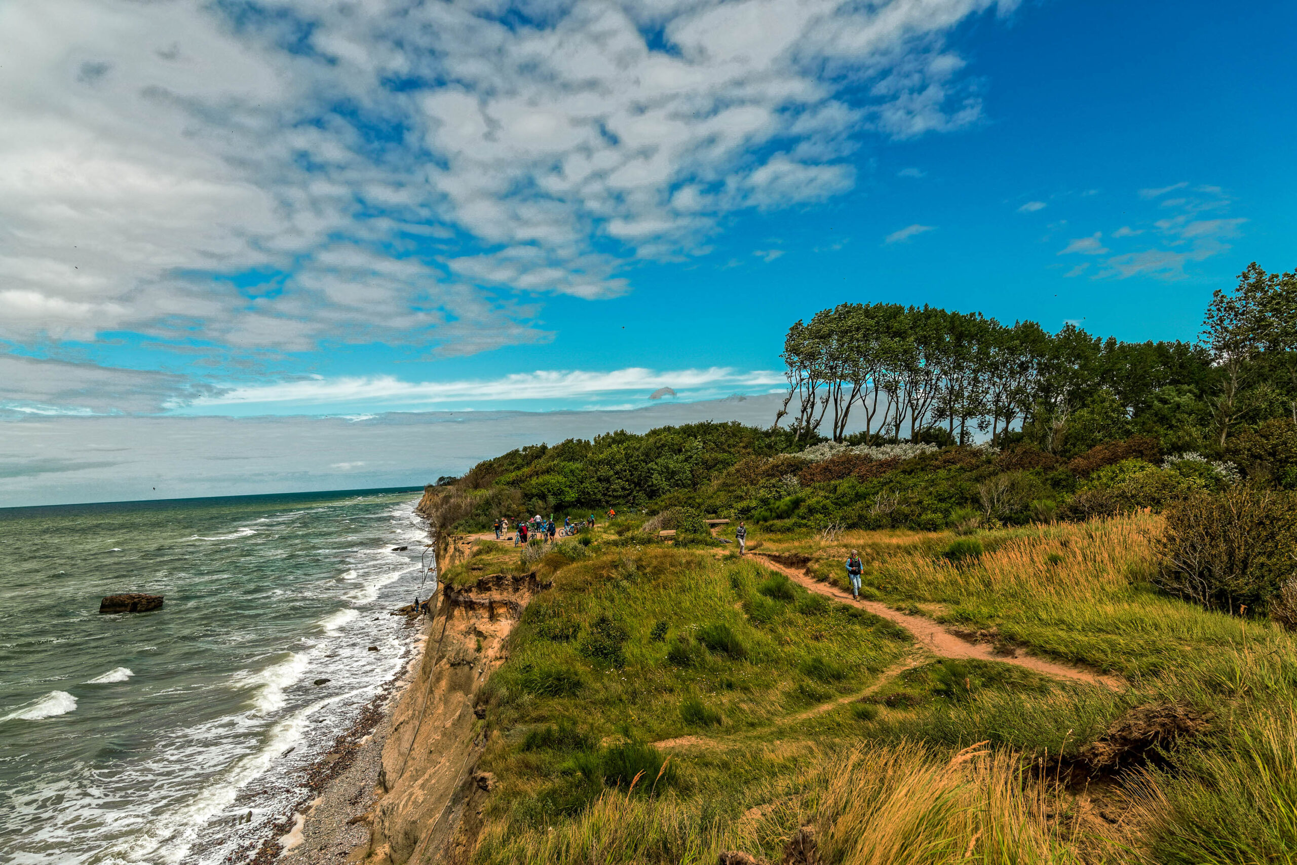 Die Aufnahme zeigt eine typische Landschaft von Fischland-Darß-Zingst, mit steilen, bewaldeten Klippen, die in die Ostsee abfallen. Der Weg deutet auf einen Wanderpfad hin, der die Besucher zu Aussichtspunkten führt. Die Personen am Rande der Klippe scheinen die Landschaft zu genießen und Fotos zu machen. Die Aufnahme fängt die Schönheit und Erhabenheit der Küstenlandschaft ein.