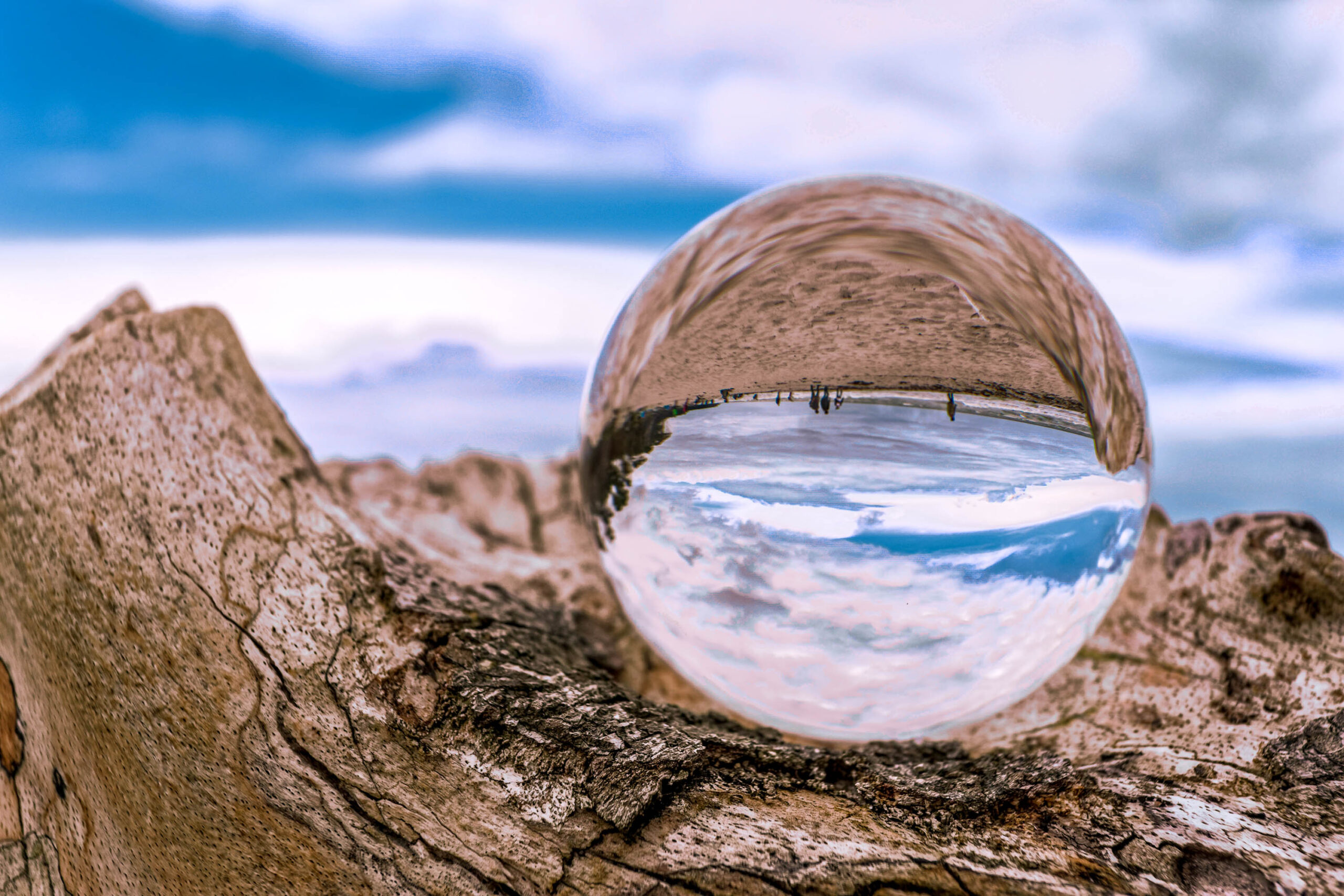 Das Bild zeigt einen Kugelreflektor, der auf einem Baumstamm platziert ist. Der Kugelreflektor spiegelt den Himmel und den Strand wider. Der Himmel ist bewölkt und der Strand ist mit Sand bedeckt. Im Hintergrund sind einige Personen zu sehen. Das Bild wurde im Juli 2020 während eines Fotosafari auf Fischland-Darss-Zingst aufgenommen.