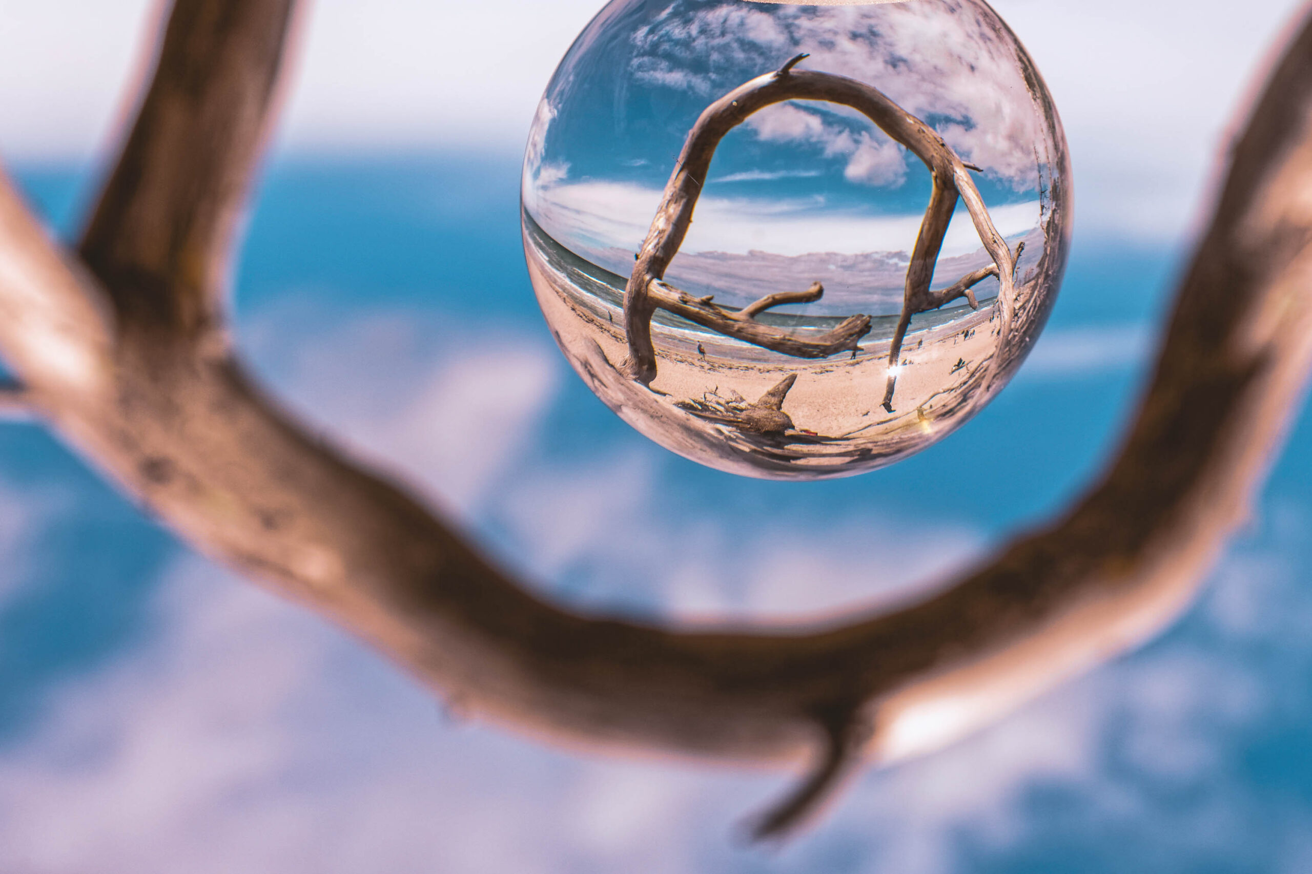 Das Bild zeigt eine ungewöhnliche Perspektive, eingefangen durch eine Kristallkugel. Im Vordergrund befindet sich ein dunkler, verzweigter Ast, der die Kugel umrahmt und einen Rahmen für die darin abgebildete Szene schafft. Durch die Kugel hindurch ist ein Strand mit feinem Sand zu sehen, der von einem ruhigen, blauen Meer und einem bewölkten Himmel begrenzt wird. Die Perspektive verzerrt die Landschaft und erzeugt einen surrealen Effekt. Die Farben sind gedämpft und wirken fast monochrom, was dem Bild eine ruhige und meditative Atmosphäre verleiht. Der Ast im Vordergrund verleiht dem Bild eine organische Note und betont die Verbindung zwischen Natur und Fotografie.