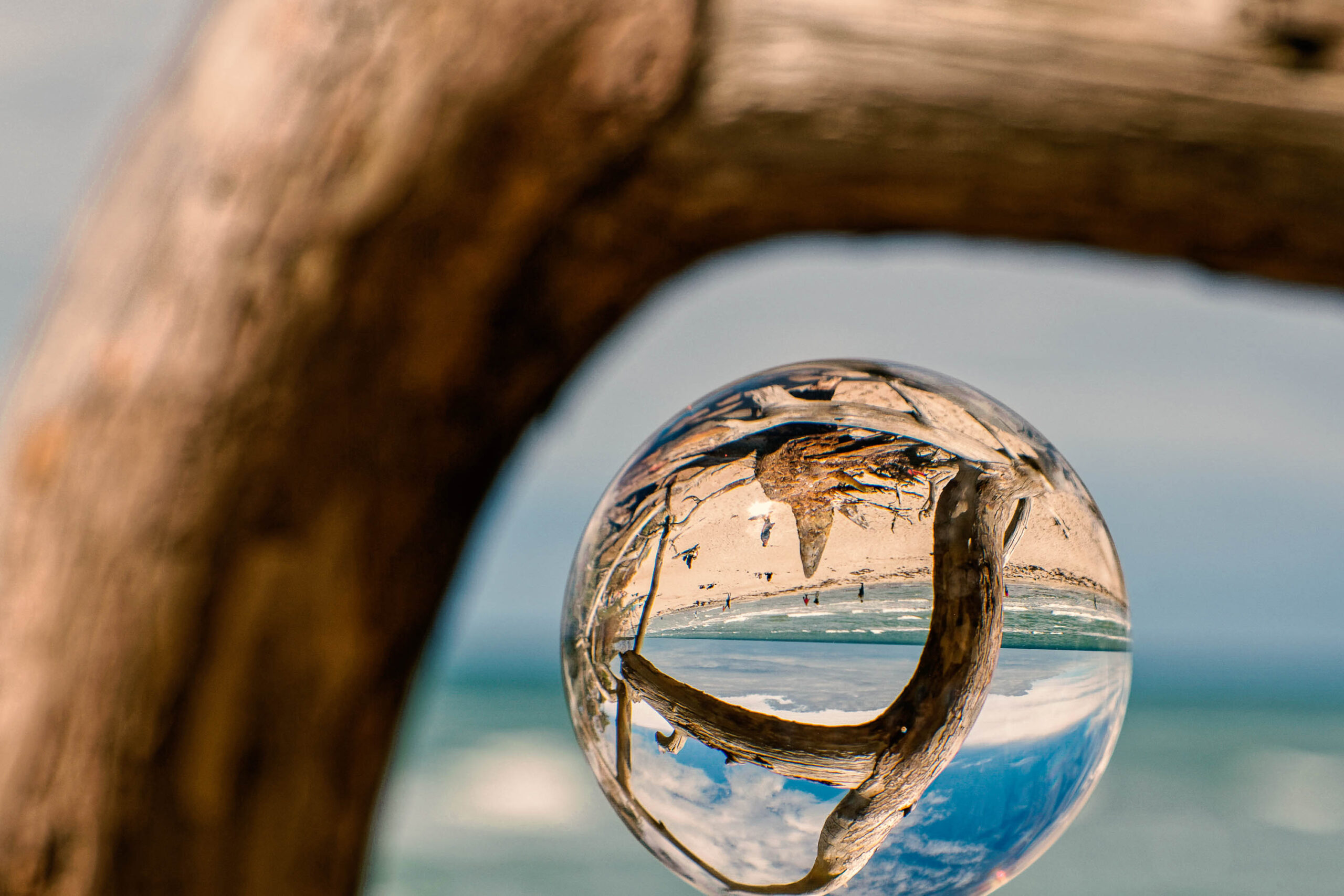 Das Bild zeigt eine Kugelreflektor-Linse, die eine verzerrte Ansicht eines Strandes einfängt. Im Vordergrund ist ein Stück Treibholz zu sehen, das durch die Linse in das Bild integriert ist. Der Strand erstreckt sich bis zum Horizont, wo er in einem blauen Himmel mit vereinzelten Wolken aufhört. Die Szene vermittelt eine friedliche und ruhige Atmosphäre. Die Aufnahme wurde im Juli 2020 während einer Fotosafari auf Fischland-Darss-Zingst aufgenommen.