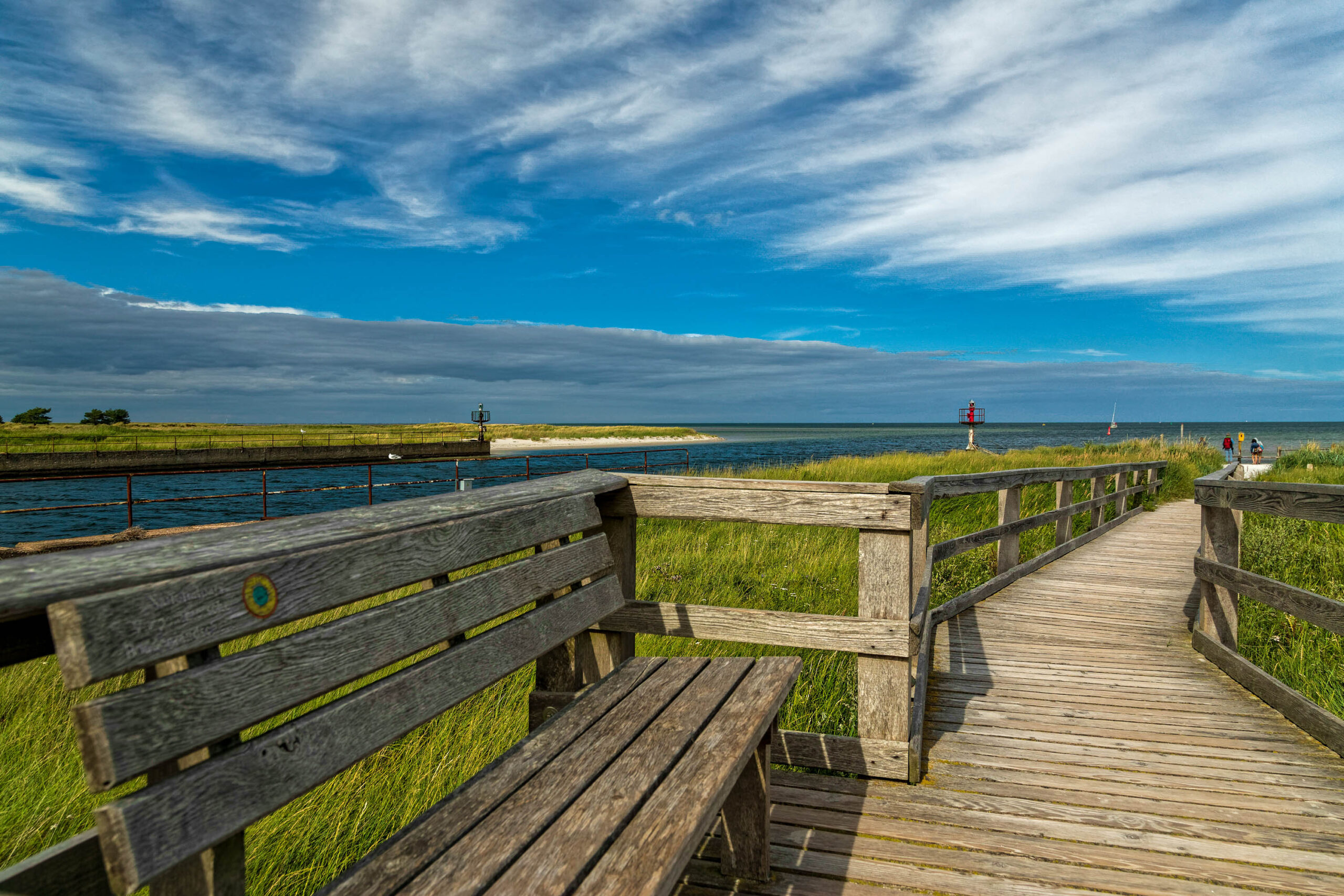 Die Aufnahme zeigt einen Holzsteg, der sich durch eine grüne Landschaft schlängelt, mit Blick auf das Ostseebad. Der Himmel ist von Wolken bedeckt, was eine dramatische Atmosphäre schafft. Im Hintergrund sind vereinzelte Personen und ein Leuchtturm zu sehen. Die Perspektive ist von einem erhöhten Standpunkt aus aufgenommen, was einen weiten Blickwinkel ermöglicht. Die Farben sind lebendig und die Schärfentiefe ist gut, was die Details der Landschaft hervorhebt. Die Aufnahme vermittelt ein Gefühl von Ruhe und Frieden.