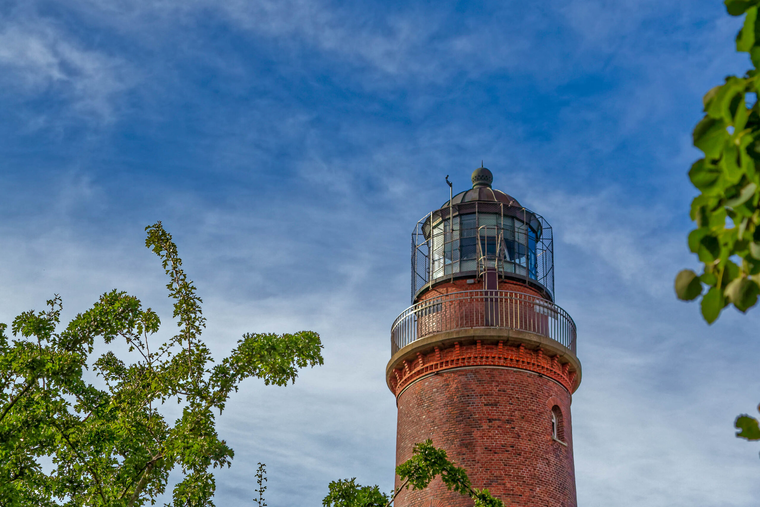Das Bild zeigt einen roten Backstein-Leuchtturm, der sich gegen einen blauen Himmel mit vereinzelten weißen Wolken erhebt. Der Leuchtturm ist von grünen Bäumen umgeben, die den unteren Teil des Turms teilweise verdecken. Die Perspektive ist von unten aufgenommen, was die Höhe des Leuchtturms betont. Die Details der Architektur des Leuchtturms sind gut erkennbar, einschließlich der Fenster, der Balkone und der Laterne auf dem Dach. Die Bäume im Vordergrund sind mit frischem, leuchtendem Grün bedeckt, was einen Kontrast zum roten Backstein des Leuchtturms bildet. Die Szene vermittelt ein Gefühl von Ruhe und Erhabenheit.