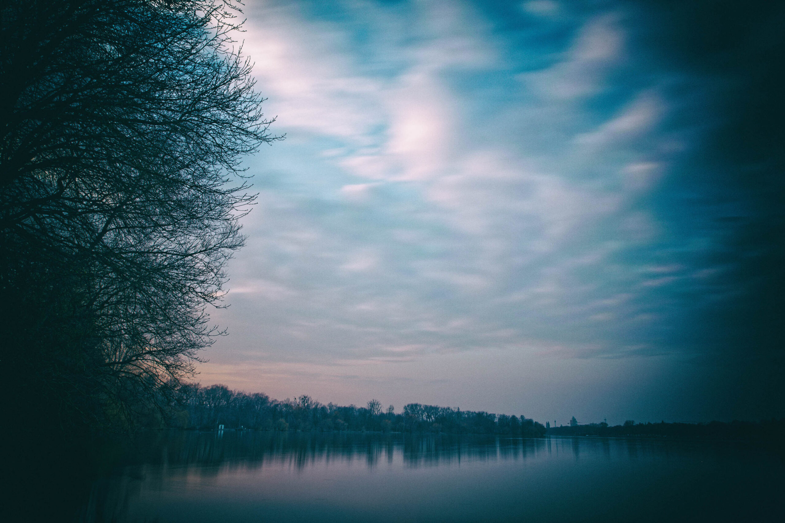 Die Aufnahme zeigt eine ruhige Seenlandschaft bei Dämmerung oder Morgendämmerung. Im Vordergrund steht ein dunkler Baum mit kahlen Ästen, der sich fast die gesamte linke Bildhälfte einnimmt. Der See spiegelt den Himmel wider, der in verschiedenen Blautönen und mit Wolkenformationen dargestellt ist. Am Horizont sind silhouettenhafte Baumreihen erkennbar. Die Farbgebung ist gedämpft und erzeugt eine ruhige, fast melancholische Stimmung. Die Aufnahme ist vermutlich in der Nähe von Hannover aufgenommen worden, möglicherweise am Maschsee oder einem anderen See in der Umgebung.