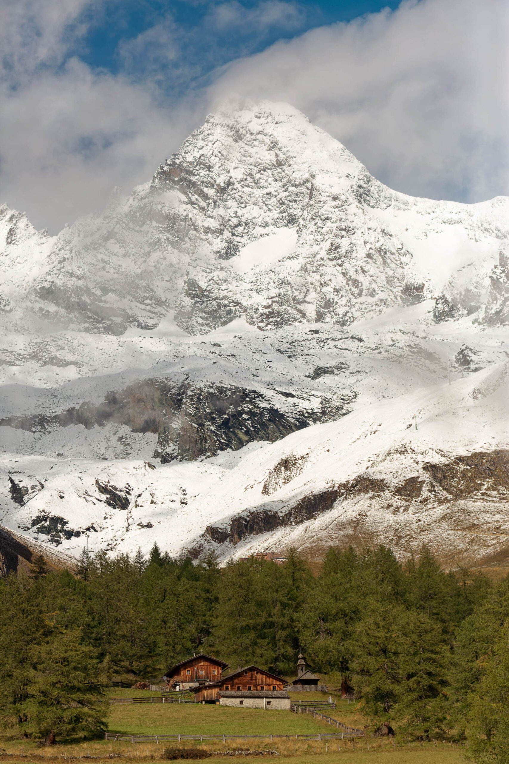 Das Foto zeigt eine traditionelle Almhütte vor dem Hintergrund eines imposanten, schneebedeckten Berges. Die Hütte ist aus Holz gebaut und hat ein steiles Satteldach. Sie ist von einem grünen Rasen umgeben, der von einer Holzzahl umgeben ist. Der Himmel ist bewölkt, aber es gibt immer noch etwas Sonnenlicht, das auf die Landschaft scheint. Die Szene vermittelt ein Gefühl von Ruhe und Frieden.