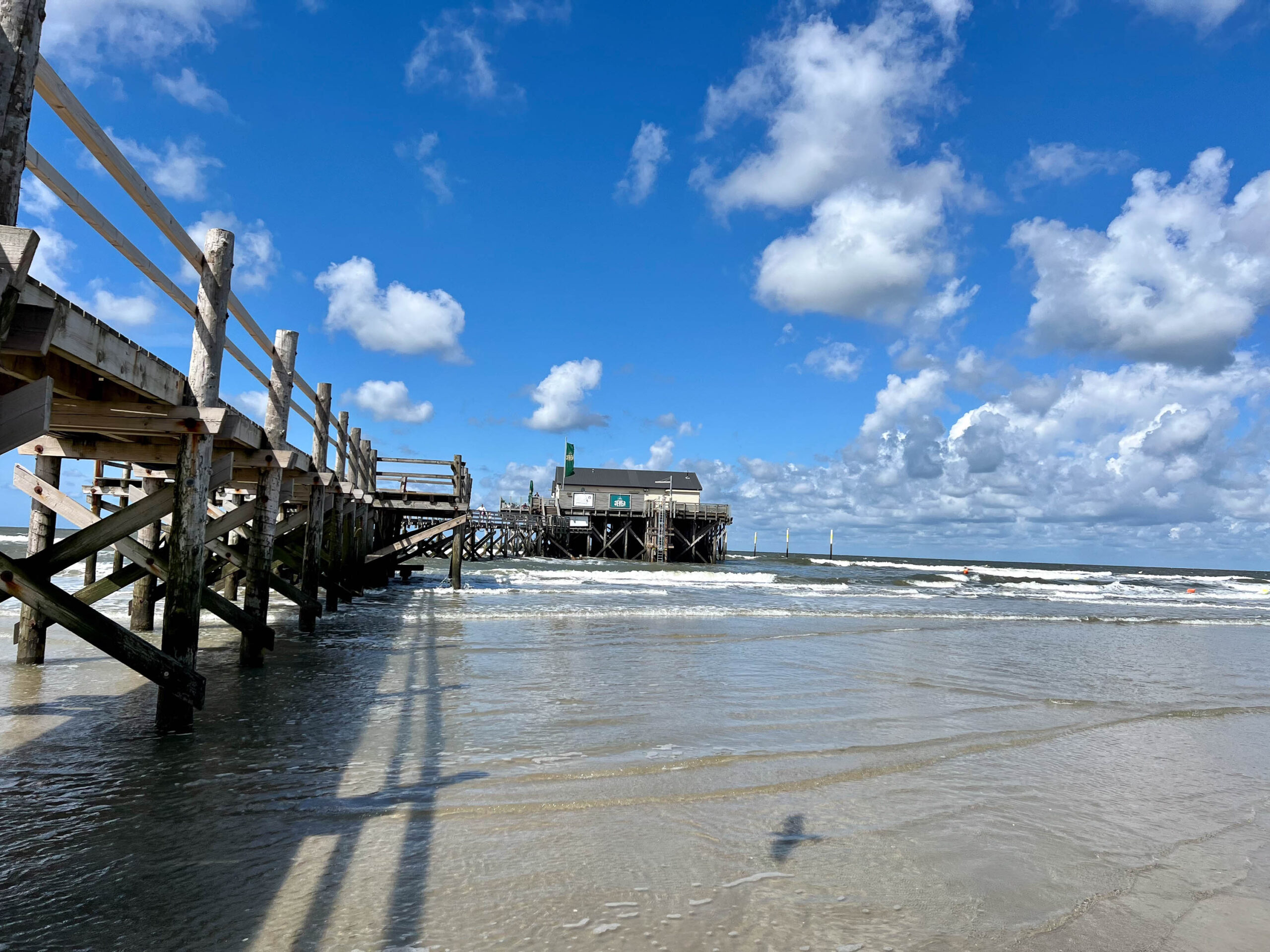 Die Aufnahme zeigt einen Pfahlbau, der sich entlang des Strandes erstreckt. Der Bau besteht aus Holzpfählen, die in den Sand gerammt sind, und darauf errichteten Gebäuden. Das Wasser des Meeres fließt sanft um die Pfähle und hinterlässt eine leichte Gischt. Der Himmel ist blau und von weißen Wolken übersät. Die Szene vermittelt eine friedliche und entspannte Atmosphäre. Die Sonne wirft lange Schatten auf den Strand.