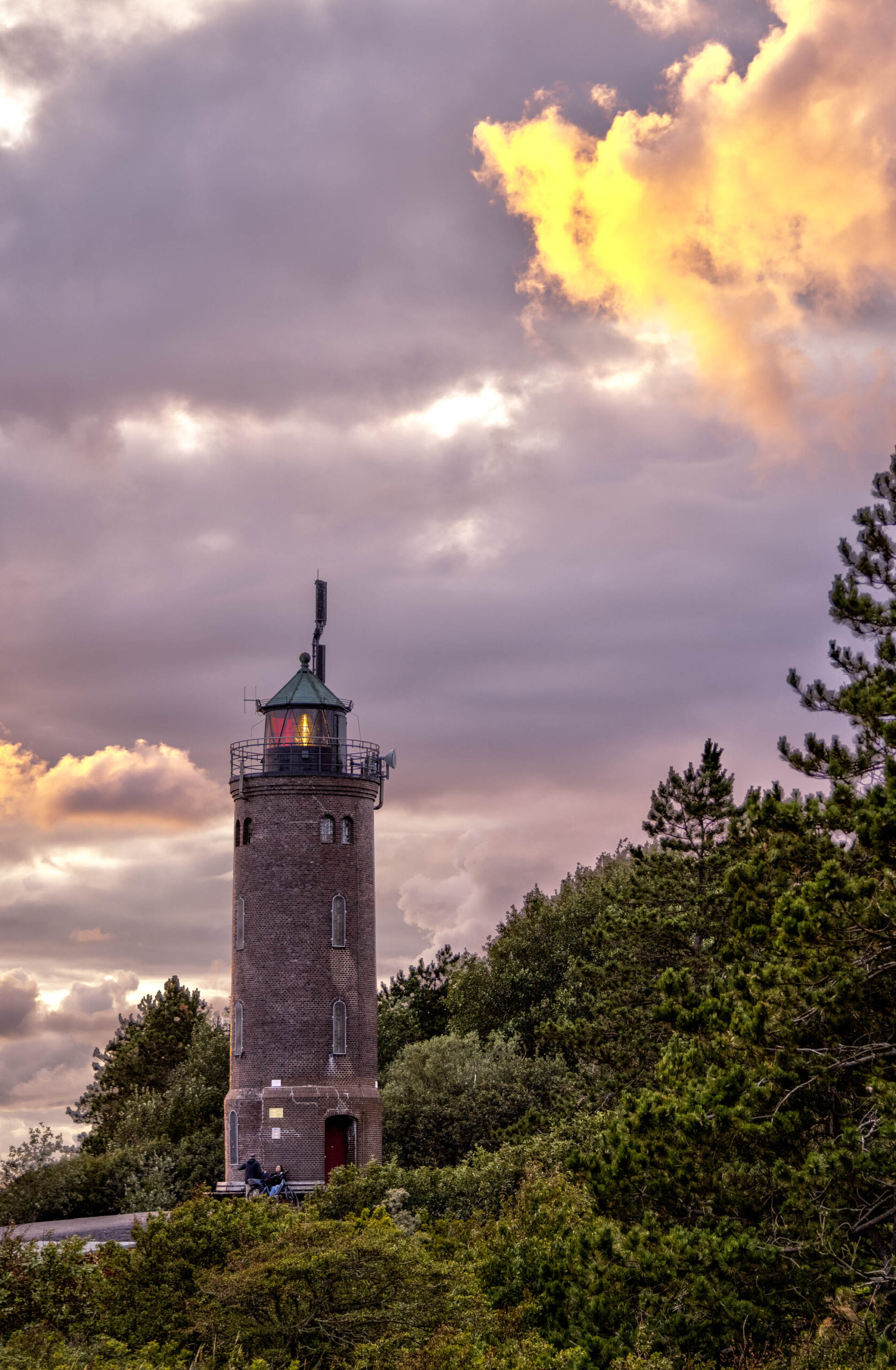 Das Bild zeigt den Böhler Leuchtturm in St. Peter-Ording bei Sonnenuntergang. Der Leuchtturm ist ein massives, zylindrisches Bauwerk aus Ziegelsteinen, das auf einem bewaldeten Hügel steht. Der Himmel ist von dramatischen Wolkenformationen in Rosa- und Violetttönen bedeckt, die das Licht der untergehenden Sonne reflektieren. Einige Personen befinden sich in der Nähe des Leuchtturms. Die Vegetation besteht hauptsächlich aus Kiefern und Sträuchern. Die Szene vermittelt eine ruhige und friedliche Atmosphäre.