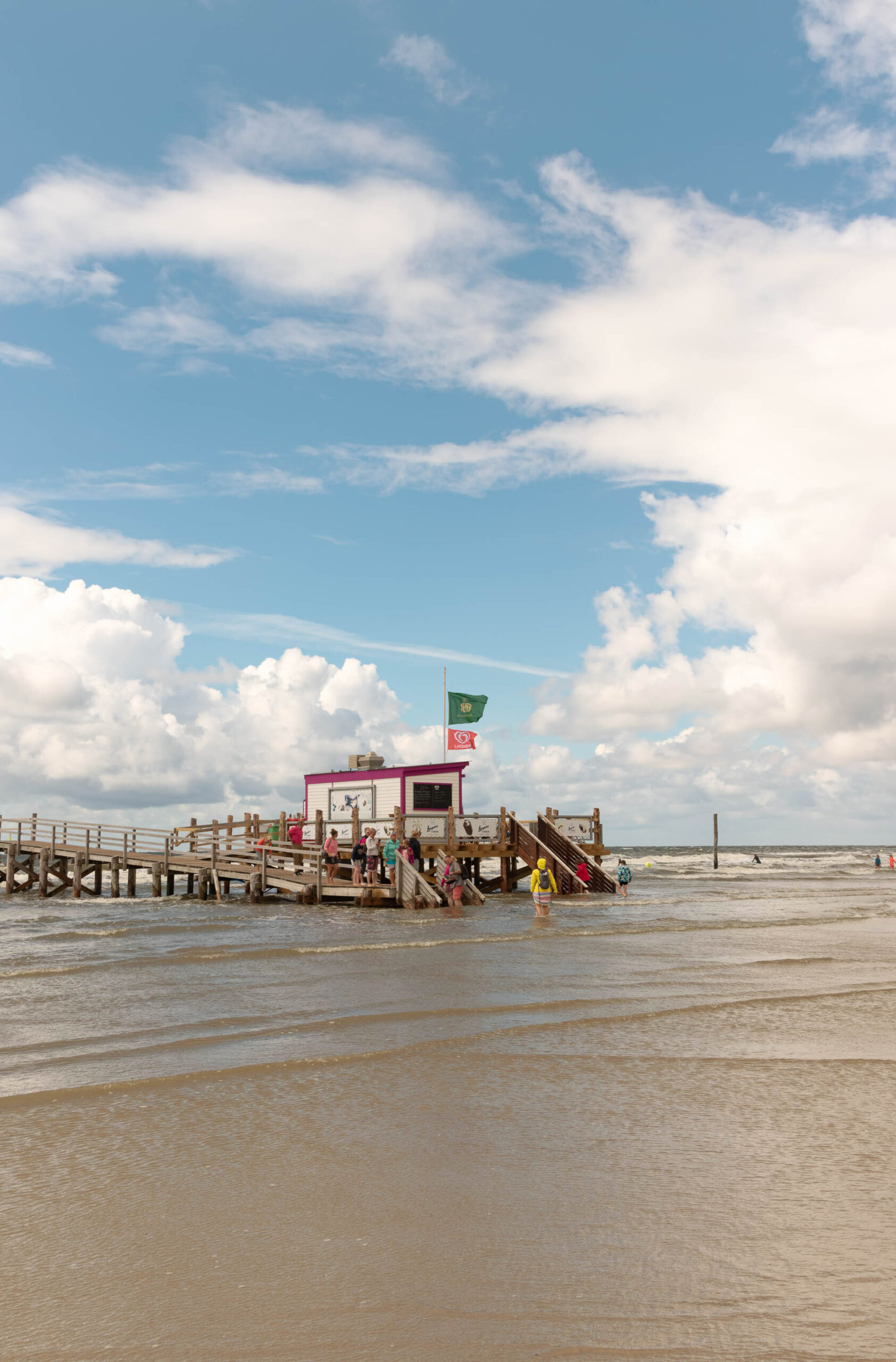 Das Bild zeigt einen Pfahlbau im Ordinger Strand bei St. Peter-Ording. Der Pfahlbau steht im flachen Wasser und ist von einer Gruppe von Menschen umgeben. Der Himmel ist blau mit weißen Wolken und das Wasser ist trüb. Im Hintergrund sind weitere Pfahlbauten und der Strand zu sehen.