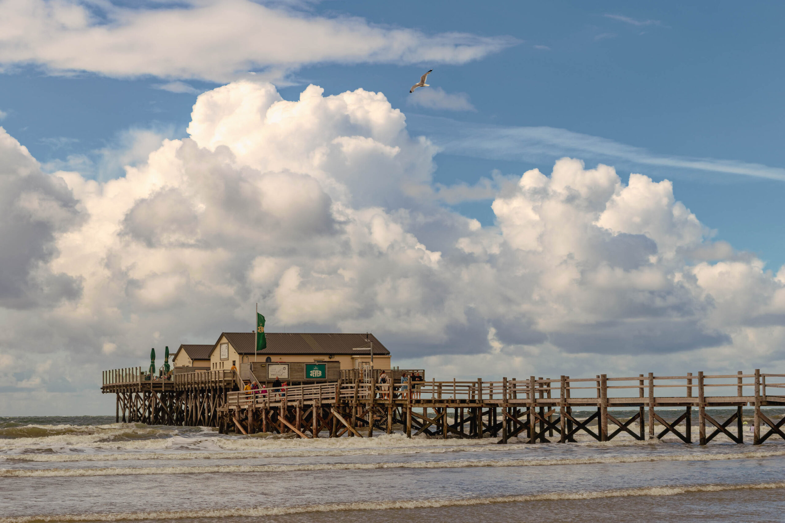 Die Aufnahme zeigt einen Pfahlbau auf dem Ordinger Strand in St. Peter-Ording. Der Pfahlbau ist ein markantes Gebäude, das auf Stelzen in der Nordsee errichtet wurde. Im Vordergrund toben Wellen, die das Ufer erreichen. Ein einzelner Vogel, vermutlich eine Möwe, fliegt am Himmel. Der Himmel ist bewölkt, aber die Sonne scheint durch die Wolken hindurch. Die Szene vermittelt eine friedliche und entspannte Atmosphäre.