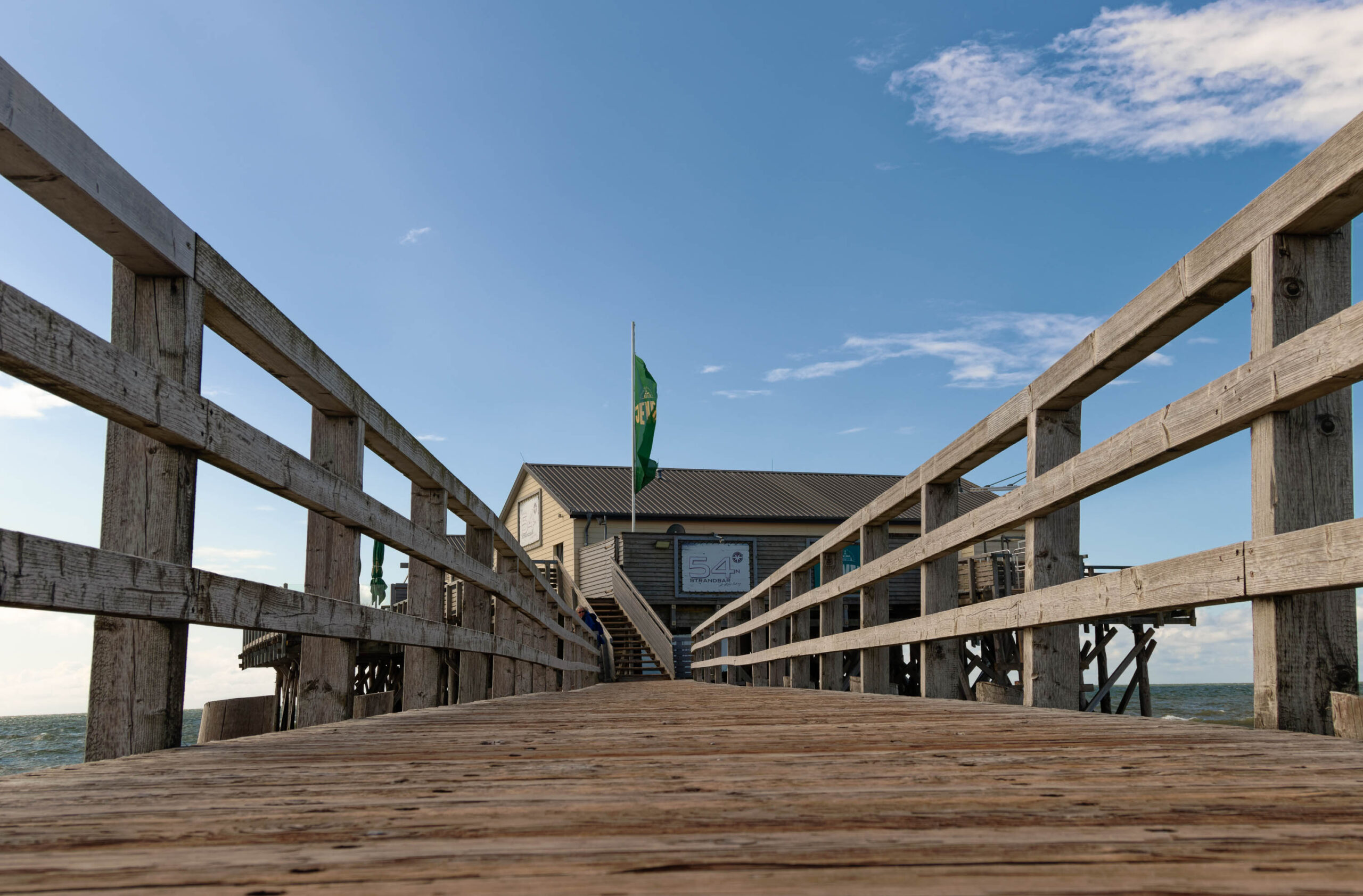 Das Foto zeigt eine Perspektive entlang eines Holzstecks auf einem Pfahlbau auf dem Ordinger Strand in St. Peter-Ording. Der Pfahlbau ist ein charakteristisches Gebäude mit einem Satteldach und einer Holzkonstruktion, die auf Stelzen in der Nordsee steht. Die Perspektive betont die Länge des Stegs und führt den Blick auf das Gebäude am Ende des Stegs. Der Himmel ist blau und wolkenlos, was auf einen sonnigen Tag hindeutet. Die Umgebung ist von Dünen und Strandflächen geprägt. Die Farben sind natürlich und die Beleuchtung ist hell.
