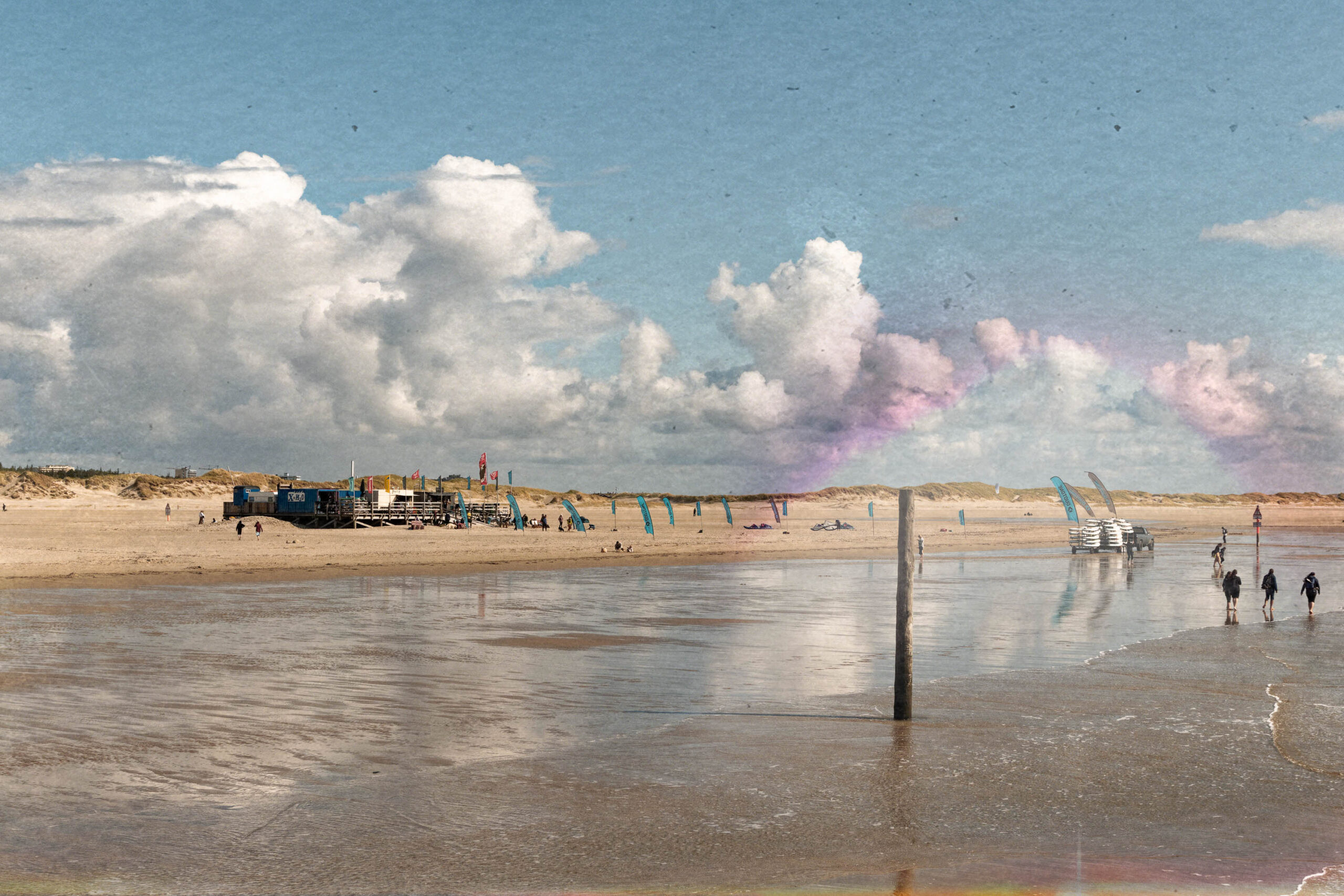 Die Aufnahme zeigt eine typische Szene vom Ordinger Strand in St. Peter-Ording. Im Vordergrund befindet sich flaches, reflektierendes Wasser, das den Himmel und die Wolken spiegelt. Im Hintergrund erhebt sich ein Pfahlbau, der aus mehreren Gebäuden besteht, die auf Stelzen errichtet sind. Diese Bauweise ist charakteristisch für die Nordseeküste und dient dazu, die Gebäude vor Überschwemmungen zu schützen.  Mehrere Personen sind auf dem Strand und in der Nähe des Pfahlbaus zu sehen. Der Himmel ist von weißen, bauschigen Wolken bedeckt, was auf eine sommerliche Atmosphäre hindeutet. Die Szene vermittelt ein Gefühl von Weite und Ruhe, typisch für die deutsche Nordseeküste.