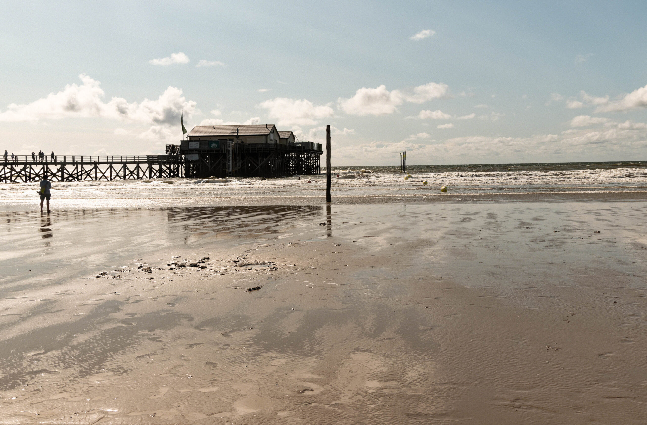 Die Aufnahme zeigt eine Szene am Ordinger Strand in St. Peter-Ording, Nordsee. Im Vordergrund erstreckt sich ein breiter Sandstrand, der durch die Ebbe freigelegt wurde und von Wellenmustern gezeichnet ist. Im Hintergrund erhebt sich ein markanter Pfahlbau, der sich über das Wasser und den Strand erstreckt. Der Himmel ist bewölkt, mit vereinzelten Sonnenstrahlen, die durch die Wolken dringen. Einige Personen sind auf dem Pfahlbau zu sehen, während eine einzelne Person am Strand steht und in Richtung des Pfahlbaus blickt. Die Szene vermittelt eine ruhige und friedliche Atmosphäre, typisch für einen Tag am Nordsee-Strand.