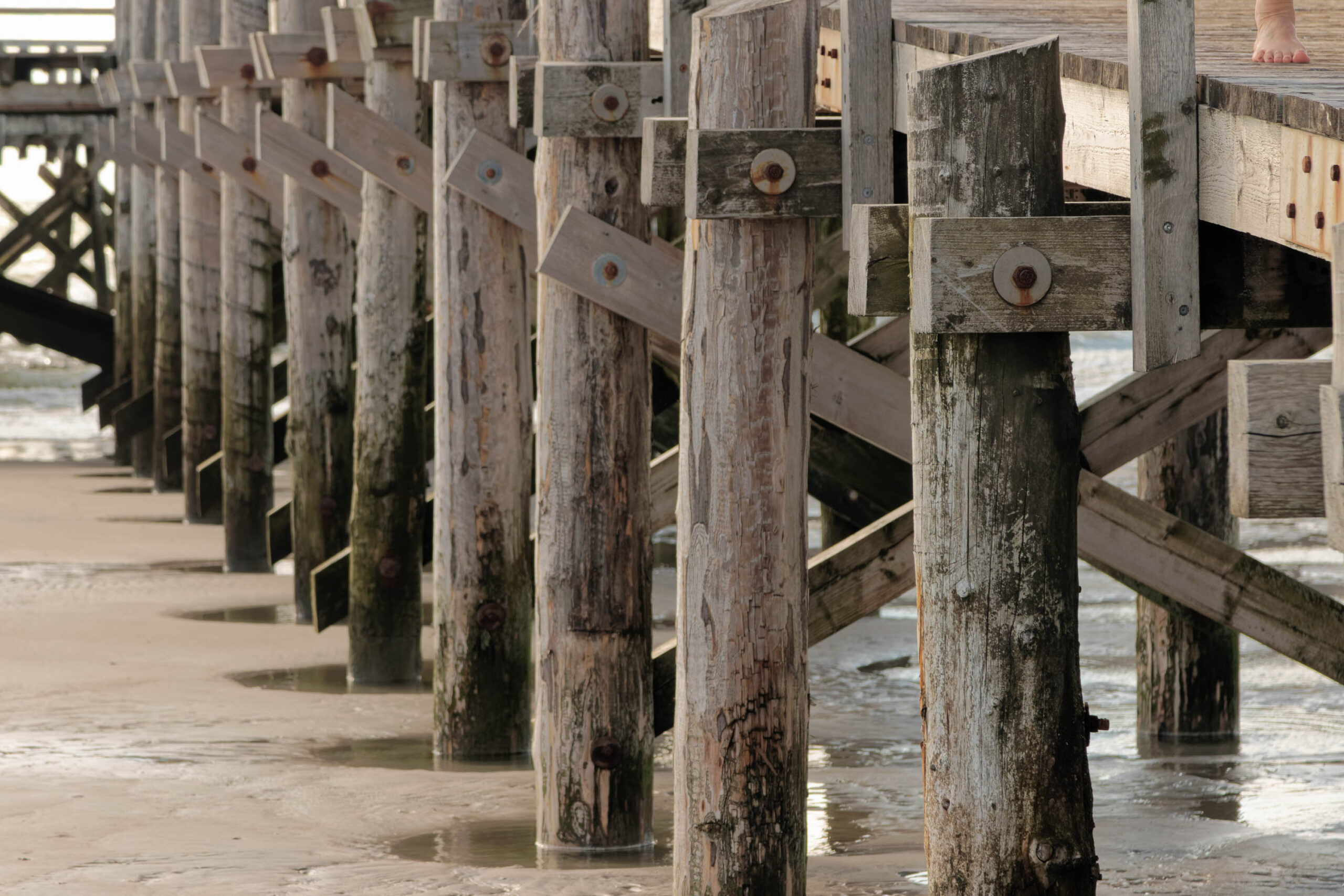 Die Aufnahme zeigt eine Detailaufnahme eines Pfahlbaus, vermutlich am Ordinger Strand in St. Peter-Ording. Die Pfähle sind aus Holz und wirken durch die Witterungseinflüsse stark verwittert. Die Textur des Holzes ist deutlich erkennbar. Im Vordergrund liegt feuchter Sand, der die Reflexionen der Pfähle und des Himmels widerspiegelt. Die Perspektive ist von unten aufgenommen, was die Höhe der Pfähle betont. Die Szene vermittelt eine ruhige und besinnliche Atmosphäre.