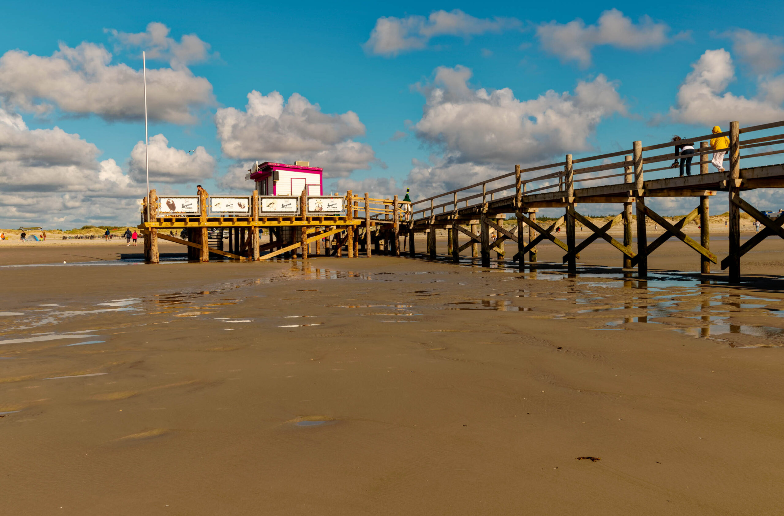 Die Aufnahme zeigt einen Pfahlbau auf dem Strand von St. Peter-Ording. Der Strand ist feucht und sandig, und der Himmel ist blau mit vereinzelten weißen Wolken. Der Pfahlbau steht auf Stelzen im Wasser und ist mit einem roten Dach versehen. Auf dem Bau befinden sich Personen. Im Hintergrund sind weitere Personen auf dem Strand zu sehen. Die Szene vermittelt eine entspannte und sonnige Atmosphäre.