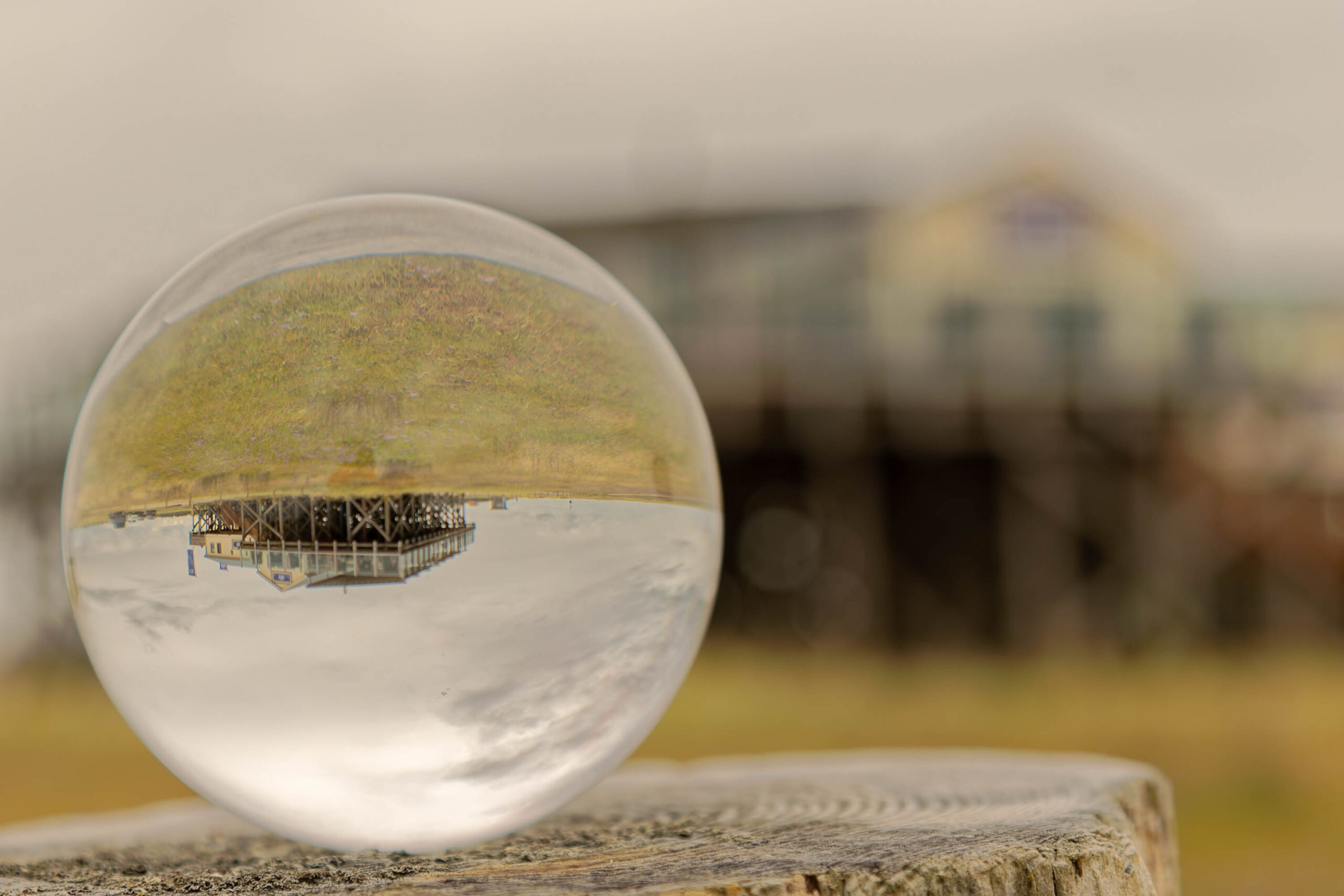 Die Fotografie zeigt eine Nahaufnahme eines Kugelreflektors, der eine verzerrte und spiegelnde Ansicht der Umgebung einfängt. Im Vordergrund befindet sich ein abgeschnittener Baumstumpf, auf dem der Kugelreflektor platziert ist. Durch die Kugel wird eine verzerrte Spiegelung des Strandes von St. Peter-Ording dargestellt, mit einem hölzernen Gebäude im Hintergrund. Der Himmel ist bedeckt und der Strand ist mit Sand bedeckt. Die Farben sind gedämpft und die Stimmung ist ruhig und friedlich.