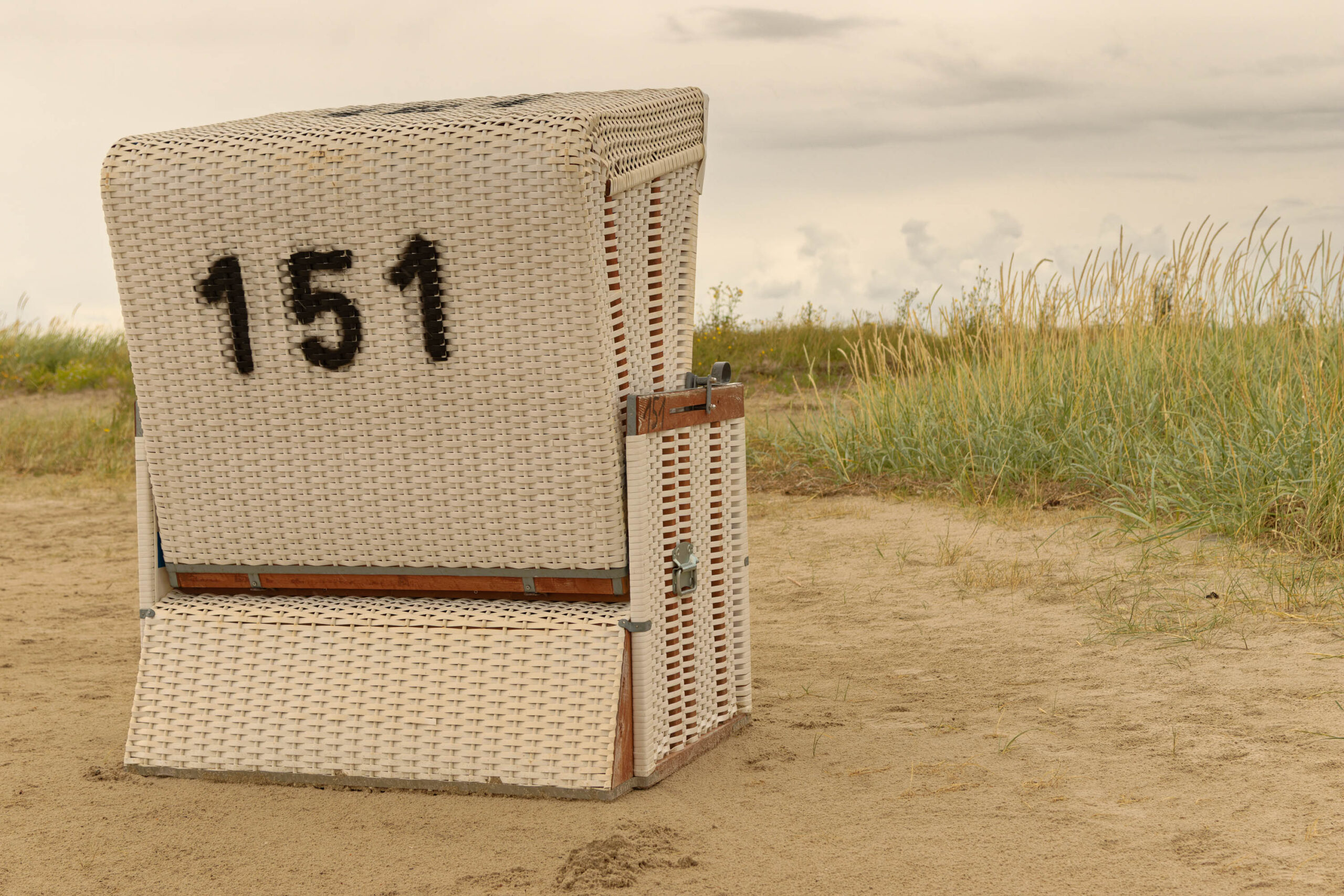 Das Bild zeigt einen einzelnen Strandkorb, der auf einem Sandstrand steht. Der Strandkorb ist aus Rattan gefertigt und mit der Nummer '151' beschriftet. Im Hintergrund sind Dünen und das Meer zu sehen. Der Himmel ist bewölkt. Das Bild wurde im Sommer 2023 in St. Peter-Ording aufgenommen.