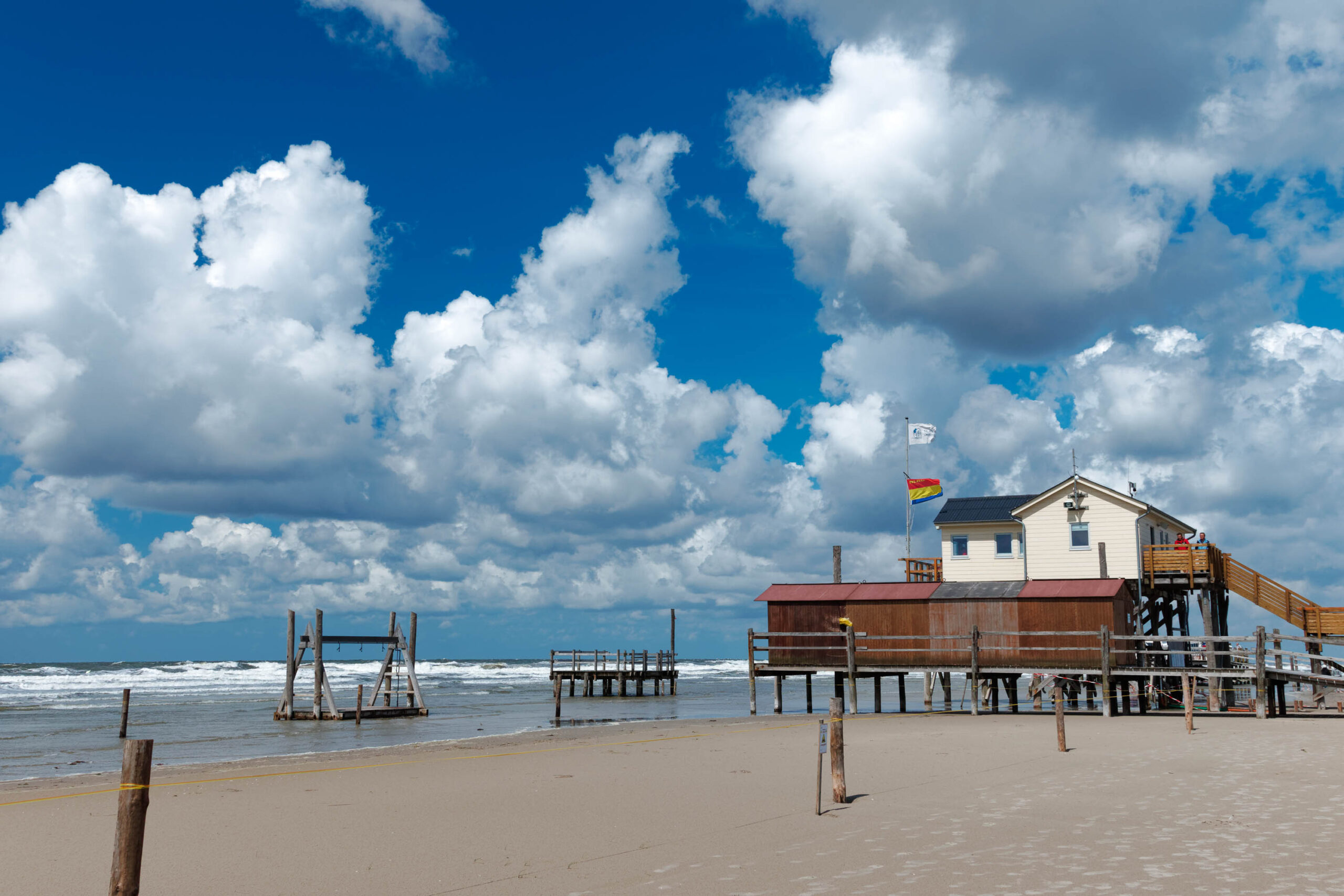 Die Aufnahme zeigt eine Szene am Ordinger Strand in St. Peter-Ording. Im Vordergrund befindet sich ein breiter Sandstrand, der von Holzpfählen und Überresten alter Pfahlbauten gesäumt ist. Im Hintergrund erhebt sich ein markanter Pfahlbau auf Stelzen, der aus einem weißen Wohnteil und einem dunkleren, erhöhten Bereich besteht. Das Gebäude steht direkt am Strand und bietet einen Blick auf das Wattenmeer und den Himmel. Der Himmel ist strahlend blau mit vereinzelten weißen Wolken, die für sommerliches Wetter sorgen. Das Bild vermittelt eine friedliche und entspannte Atmosphäre.