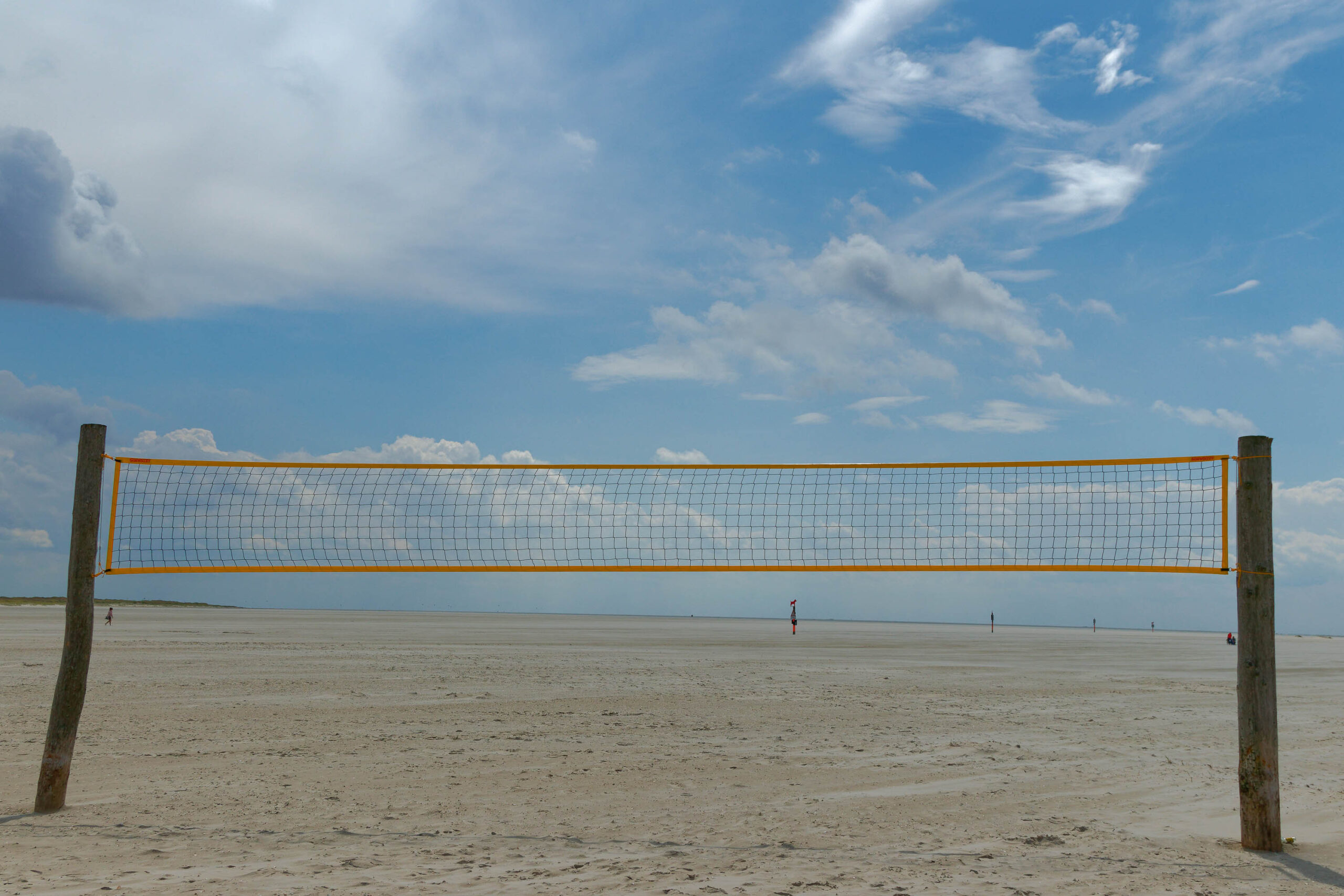 Das Bild zeigt ein Volleyballnetz, das auf dem weitläufigen Sandstrand von St. Peter-Ording aufgestellt ist. Die Pfahlbauten, die das Netz tragen, sind aus Holz und ragen aus dem Sand. Der Himmel ist bewölkt, mit hellen Wolken, die das Licht reflektieren. Im Hintergrund sind Personen zu sehen, die sich auf dem Strand bewegen. Die Szene vermittelt eine friedliche und entspannte Atmosphäre, typisch für einen Sommertag an der Nordsee.