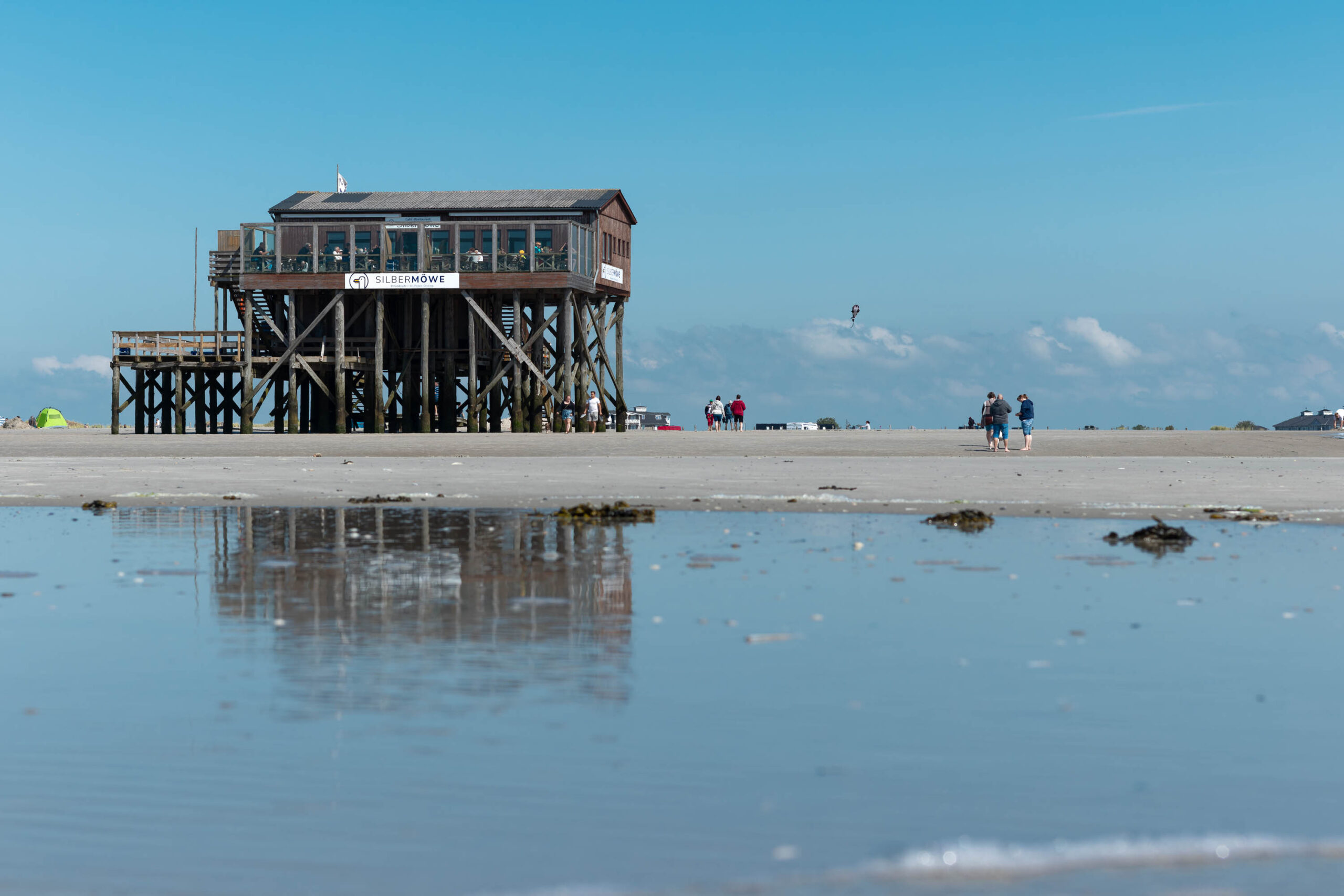 Das Bild zeigt einen Pfahlbau, der auf Stelzen in flachem Wasser steht. Es ist ein sonniger Tag, und der Himmel ist blau. Der Strand ist weitläufig und sandig. Im Hintergrund sind einige Personen zu sehen, die sich auf dem Strand aufhalten. Das Gebäude ist das Restaurant Silbermöwe in St. Peter-Ording.