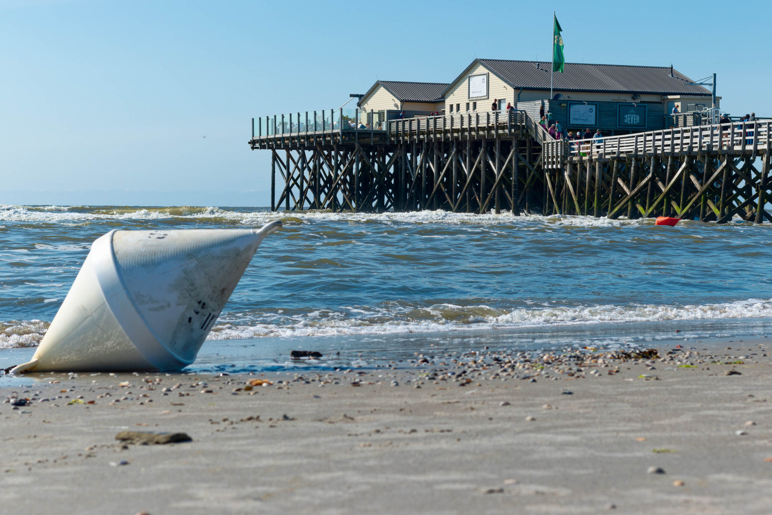 Das Bild zeigt einen Strandabschnitt mit einem weißen Boje im Vordergrund. Der Boje liegt teilweise im Sand und wird von kleinen Wellen berührt. Im Hintergrund erhebt sich ein Pfahlbau, der auf Stelzen in der Nordsee steht. Der Himmel ist blau und sonnig, was auf einen schönen Sommertag hindeutet. Menschen sind auf dem Pfahlbau zu sehen, die die Aussicht genießen. Der Strand ist mit Sand und einigen Treibholzstücken bedeckt. Die Szene vermittelt eine entspannte und friedliche Atmosphäre.