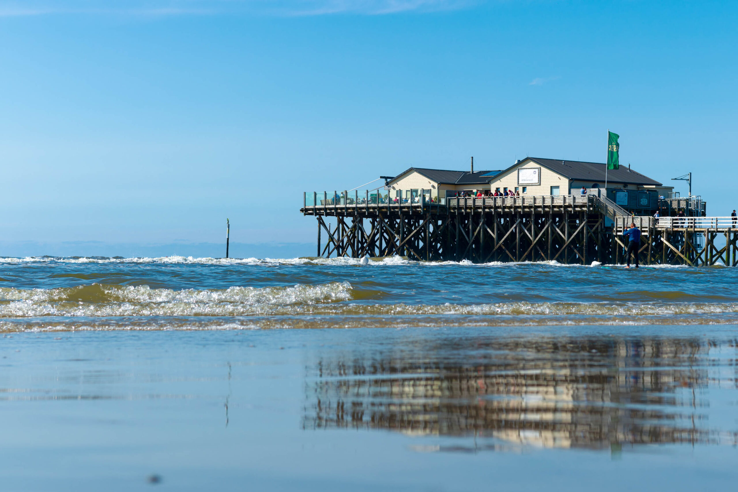 Das Bild zeigt einen Pfahlbau, der auf Stelzen in der Nordsee steht. Der Bau ist aus Holz und hat mehrere Stockwerke. Auf den Terrassen des Pfahlbaus befinden sich Personen. Im Vordergrund ist der Strand bei Ebbe zu sehen, mit feuchtem Sand und kleinen Wellen. Der Himmel ist blau und wolkenlos. Die Szene vermittelt eine ruhige und entspannte Atmosphäre.