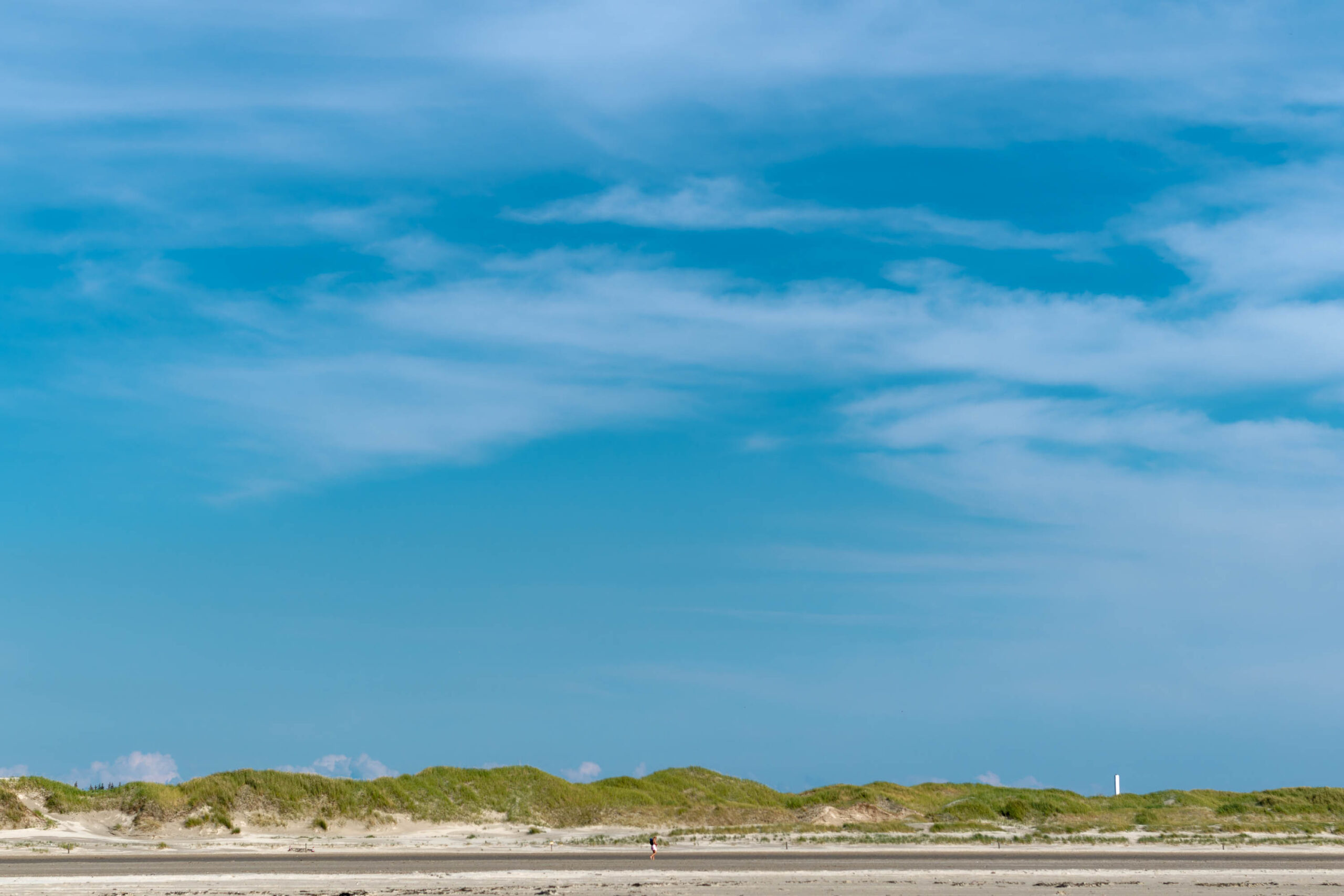 Die Aufnahme zeigt eine weite Küstenlandschaft, dominiert von einem hellen Sandstrand und einem Horizont aus grünen Dünen. Der Himmel ist strahlend blau mit vereinzelten, weißen Wolken. Im Hintergrund ist ein Pfahlbau erkennbar, der typisch für die Küste von St. Peter-Ording ist. Der Strand ist weitläufig und fast menschenleer, was eine friedliche und ruhige Atmosphäre schafft. Die Dünen sind dicht bewachsen und bilden einen schönen Kontrast zum hellen Sand.