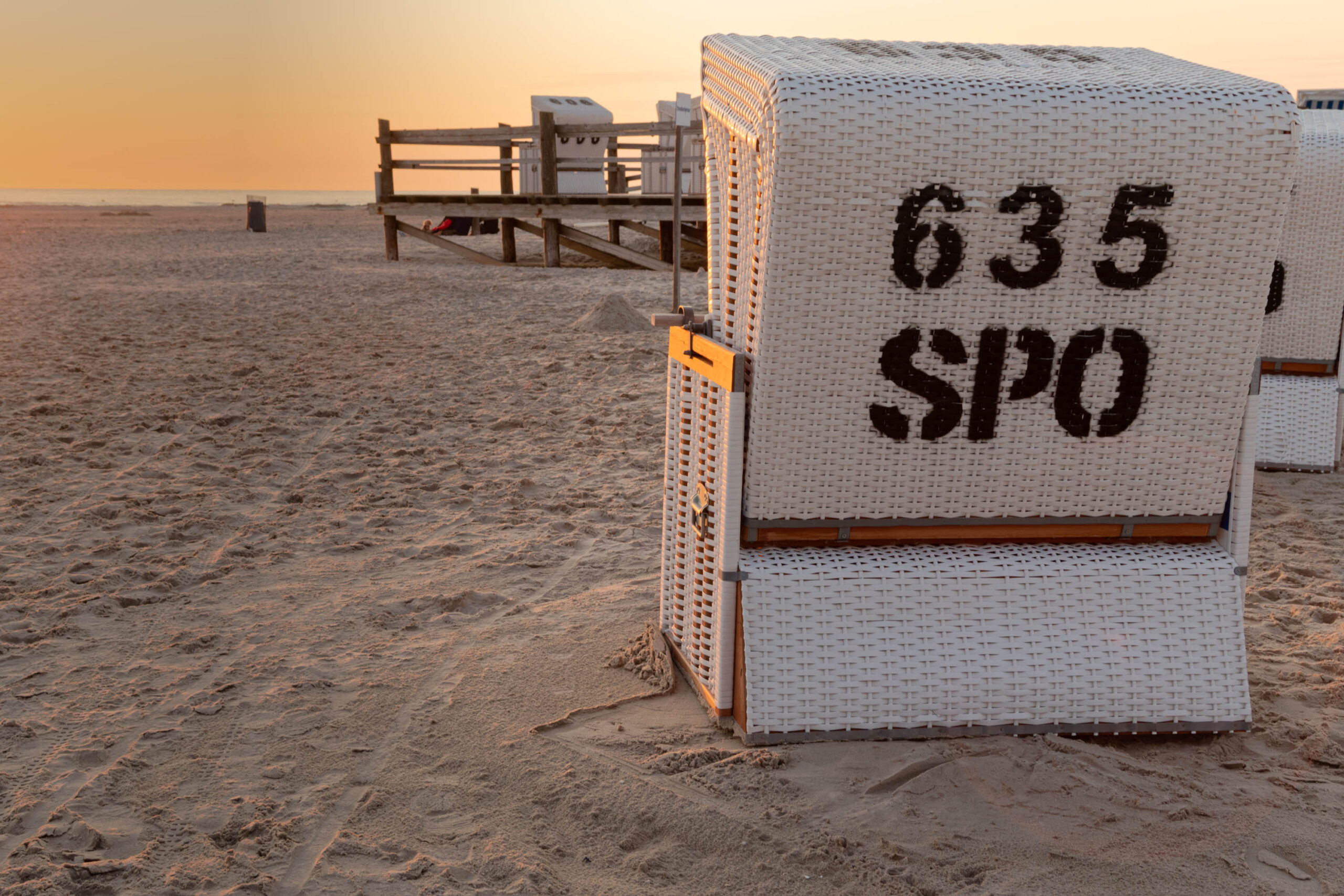 Das Bild zeigt einen weißen Strandkorb, der auf einem Sandstrand steht. Auf dem Korb sind die Nummern '635' und 'St. Peter-Ording' (kurz SPO) aufgedruckt. Im Hintergrund befindet sich ein Pfahlbau, der typisch für die Küste von St. Peter-Ording ist. Der Himmel ist in warmen Farben des Sonnenuntergangs gehalten. Der Sand ist feucht und reflektiert das Licht. Die Szene vermittelt eine friedliche und entspannte Atmosphäre.