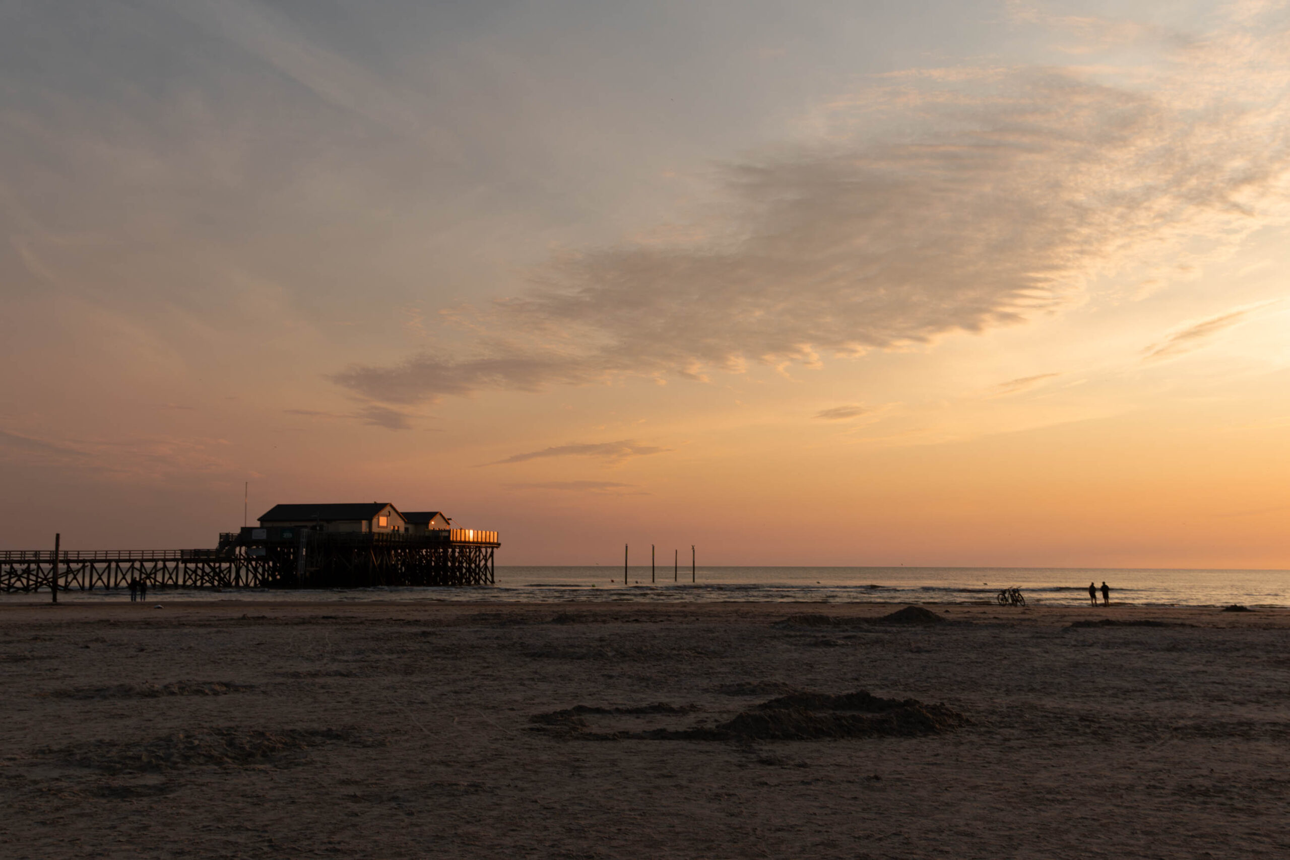 Die Aufnahme zeigt einen Pfahlbau, der sich am Strand von St. Peter-Ording erstreckt. Der Himmel ist in warmen Orange- und Rottönen gehalten, was auf einen Sonnenuntergang hindeutet. Der Strand ist weitläufig und sandig, mit vereinzelten Dünen im Vordergrund. Einige Personen sind am Strand zu sehen, die den Sonnenuntergang genießen. Die Szene vermittelt eine ruhige und friedliche Atmosphäre.