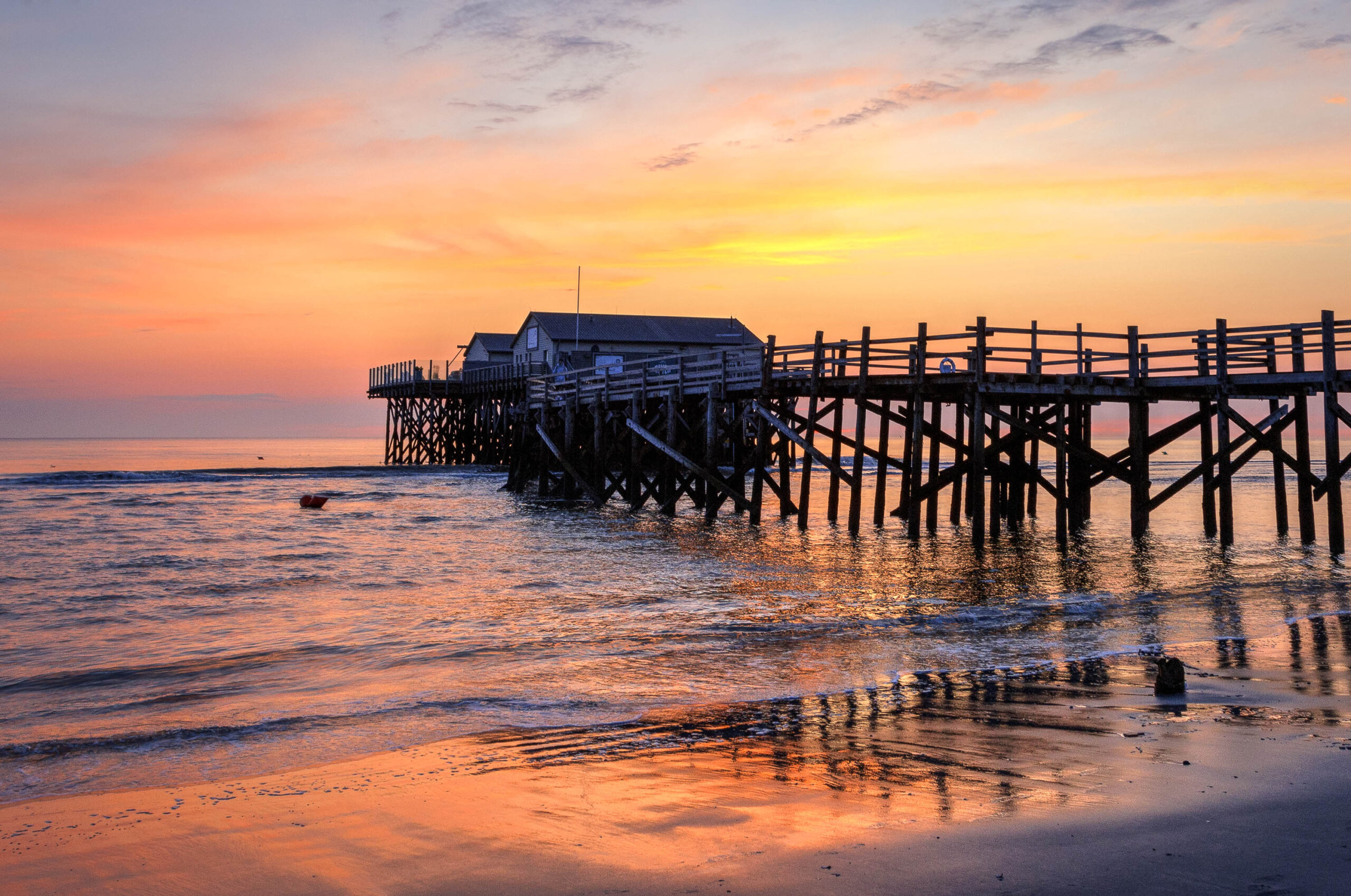 Das Bild zeigt einen Pfahlbau, der sich über das Wasser des Ordinger Strandes erstreckt. Der Himmel ist in warmen Farben des Sonnenuntergangs gehalten, die sich im Wasser spiegeln. Der Pfahlbau besteht aus mehreren Gebäuden mit schiefen Dächern und ist auf Pfählen errichtet, die in den Meeresboden getaucht sind. Der Strand im Vordergrund ist sandig und leicht feucht. Einige Treibholzstücke liegen am Strand. Die Szene vermittelt eine friedliche und ruhige Atmosphäre.