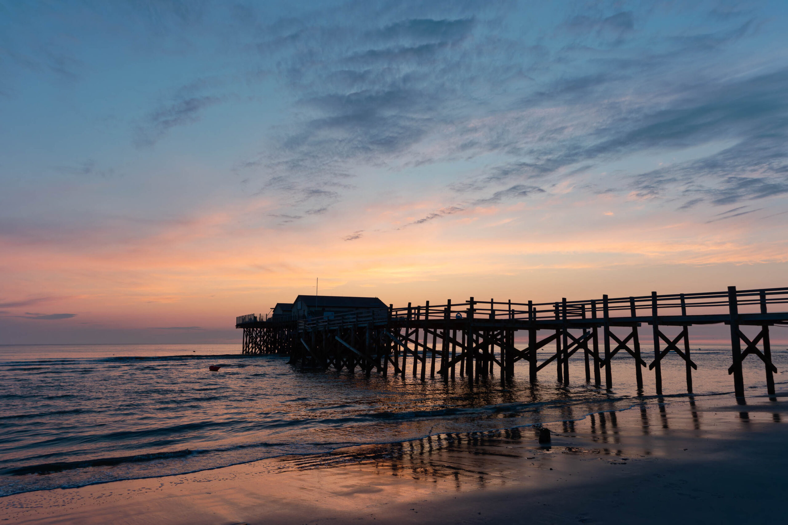 Das Bild zeigt einen Pfahlbau, der sich in das Meer hinein erstreckt. Der Himmel ist in warmen Farben wie Orange, Rosa und Blau gemalt, was auf einen Sonnenuntergang hindeutet. Der Strand ist dunkel und spiegelt die Farben des Himmels wider. Einige Personen sind auf dem Pfahlbau zu sehen. Die Szene vermittelt eine ruhige und friedliche Atmosphäre.