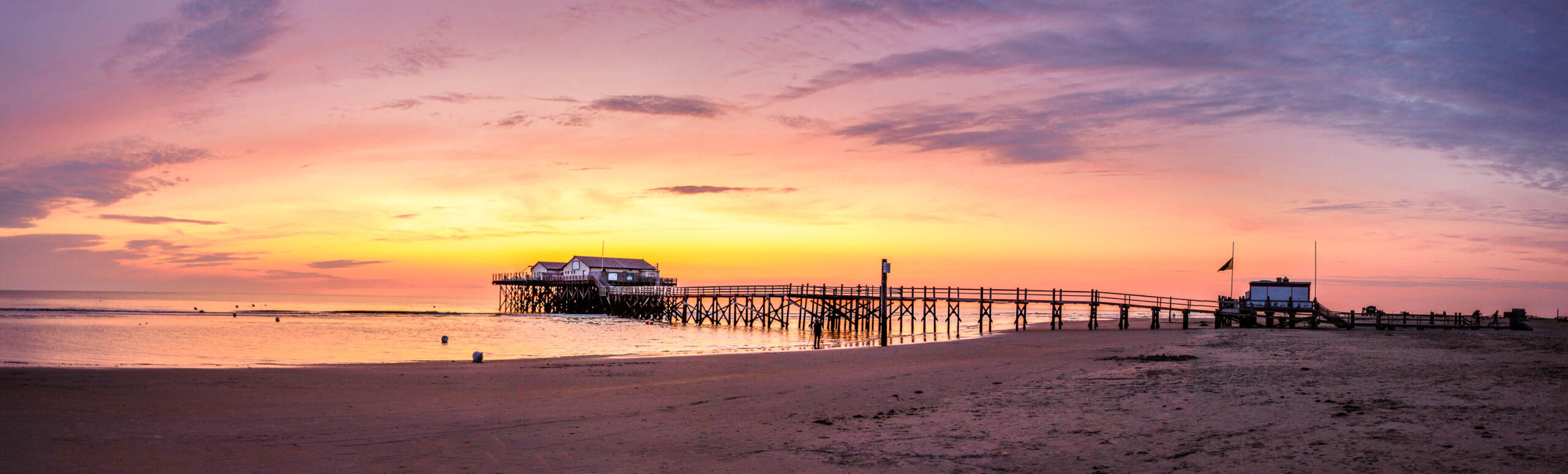 Das Bild zeigt einen Pfahlbau auf dem Ordinger Strand bei St. Peter-Ording. Der Pfahlbau steht auf Stelzen im flachen Wasser und ist von einem langen Holzsteg verbunden, der sich über den Strand erstreckt. Im Hintergrund ist ein weiterer, kleinerer Pfahlbau sowie ein Rettungsschwimmerstand zu sehen. Der Himmel ist in leuchtenden Farben des Sonnenuntergangs gehalten, hauptsächlich in Rosa, Orange und Violett. Der Strand ist feucht und reflektiert das Licht des Himmels. Einige Personen sind im Hintergrund zu erkennen, die den Sonnenuntergang genießen. Die Szene vermittelt eine friedliche und entspannte Atmosphäre.