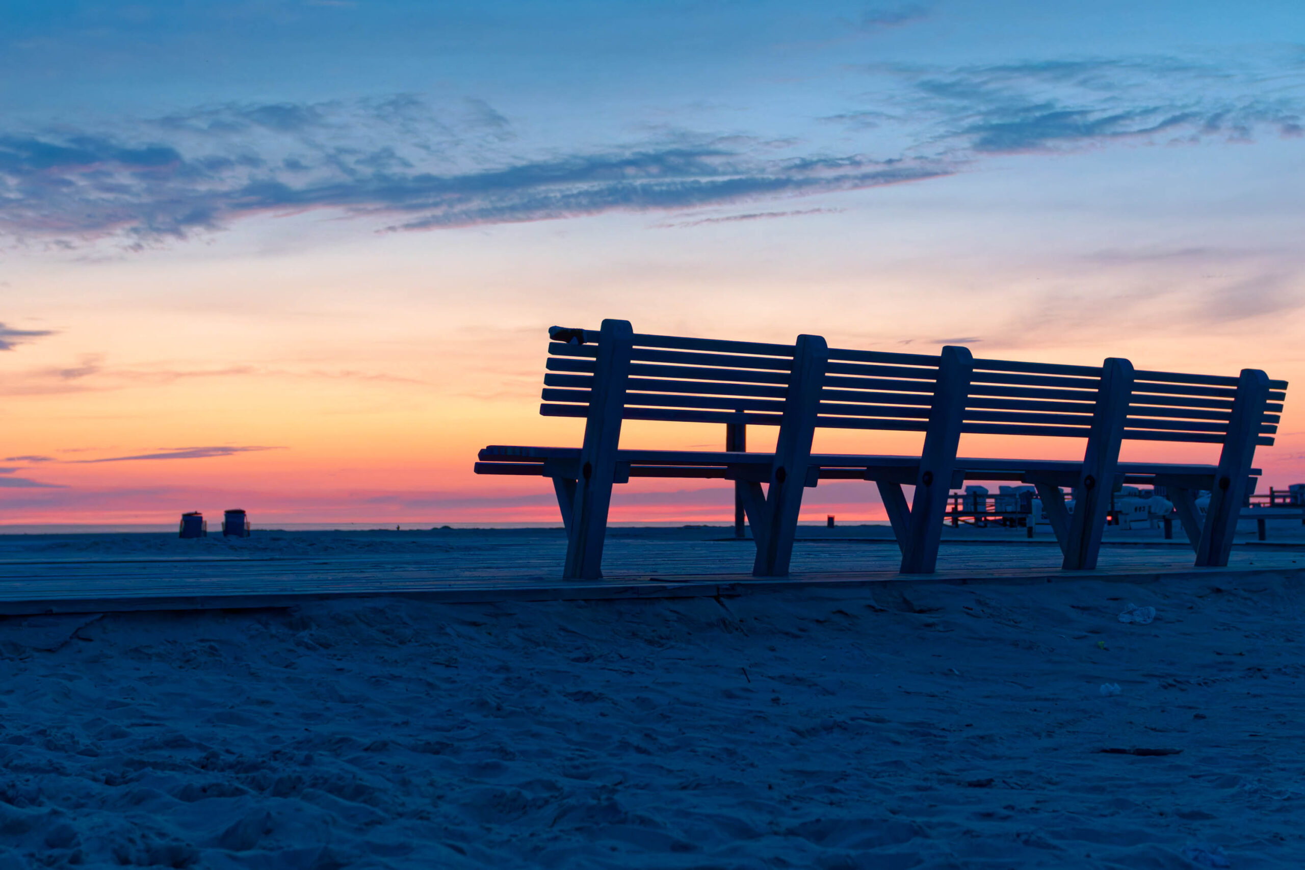 Das Foto zeigt eine Bank auf Stelzen (Pfahlbau) am Strand von St. Peter-Ording. Der Himmel ist in warmen Farben (rosa, orange) gefärbt, was auf einen Sonnenuntergang hindeutet. Der Strand ist sandig und weitläufig. Im Hintergrund sind weitere Pfahlbauten und eine Küstenlinie erkennbar. Die Szene vermittelt eine ruhige und friedliche Atmosphäre.